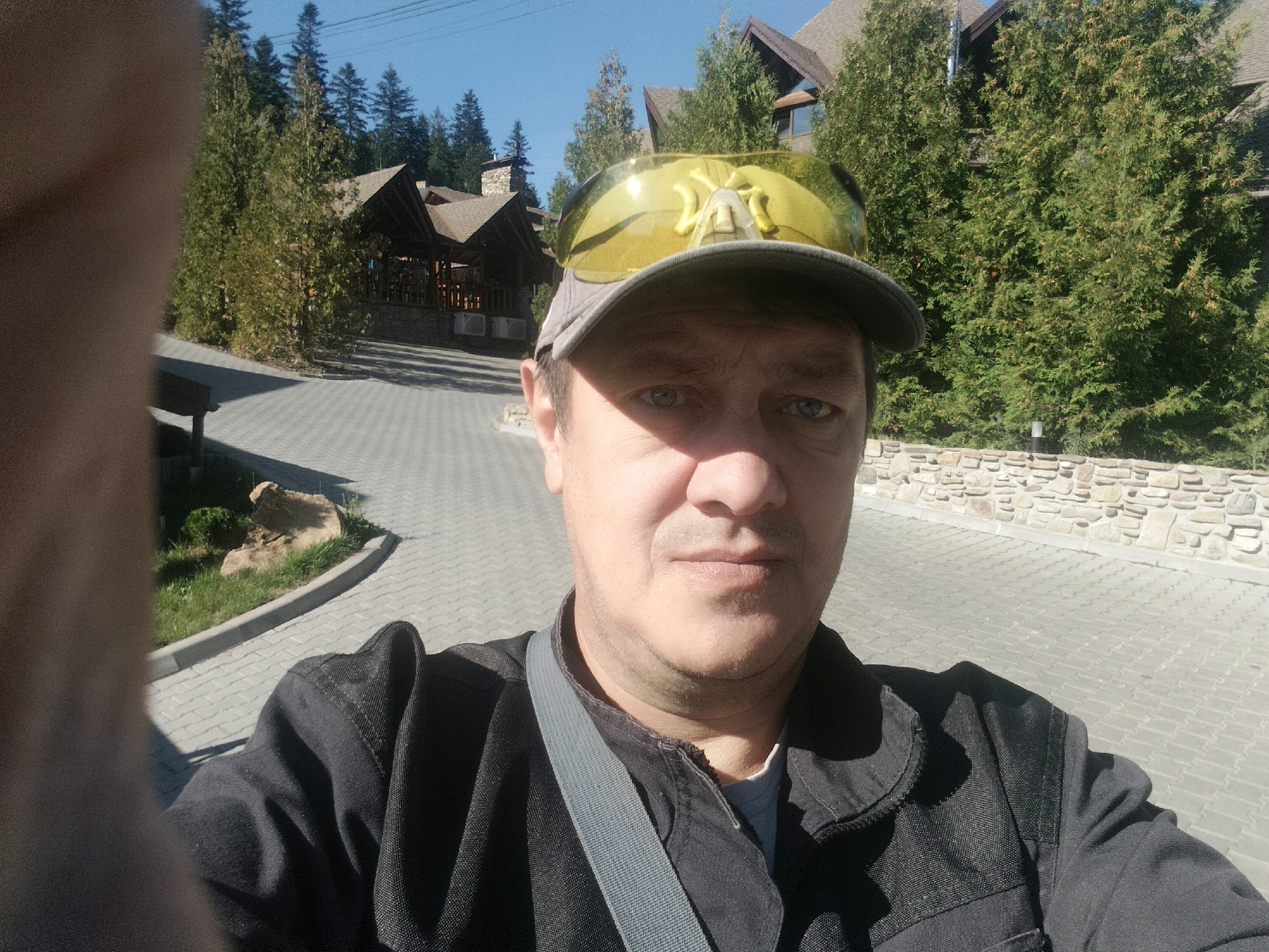 A man is taking a selfie outdoors in front of a building with a pitched roof, surrounded by trees. He is wearing a grey baseball cap and yellow sunglasses. The ground is paved with grey bricks. The man's left hand is partially visible in the bottom left corner of the frame. A grey strap is visible across his chest. The building has a stone wall and appears to be a lodge or resort. The sky is clear and blue. The image appears to have been taken during daylight hours.