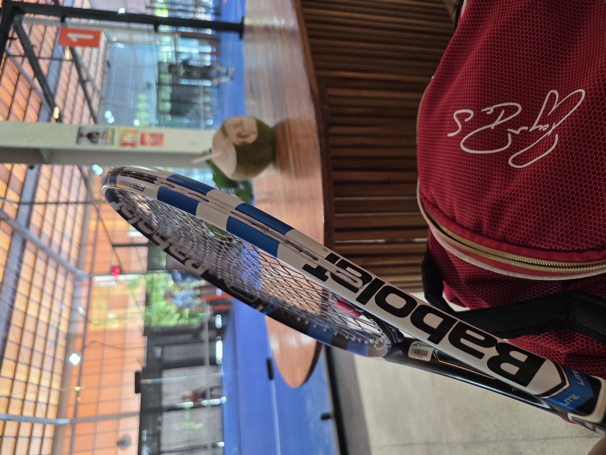 The image shows a Babolat tennis racket leaning against a red bag with a white signature on it. A coconut with a straw sits on a wooden surface in the background. The setting appears to be an indoor public space, possibly a shopping mall or airport terminal, with a view of the building's interior architecture visible through the glass windows in the background.