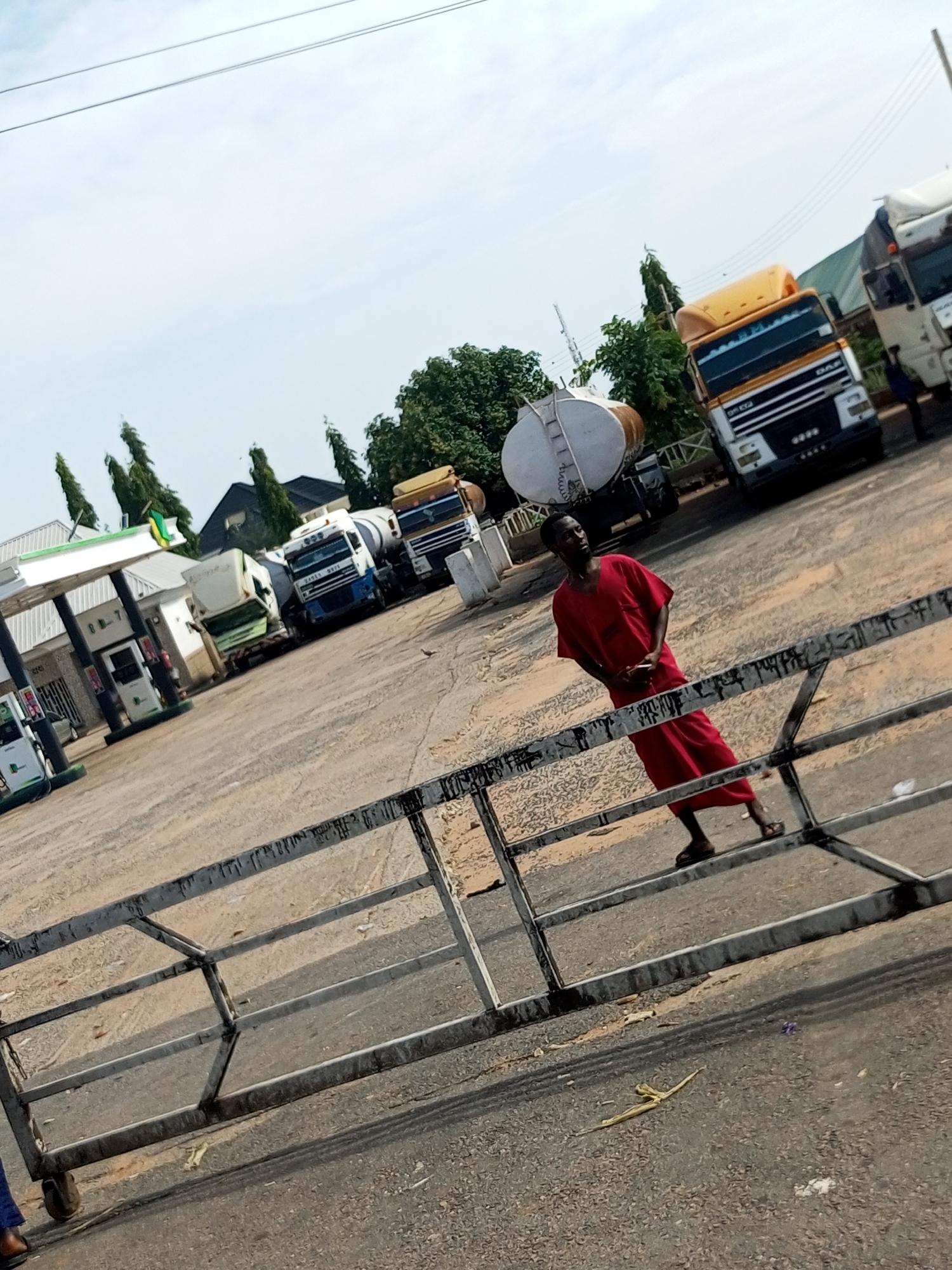 A man in a red robe stands at a gas station in Tumfure, Nigeria. He is holding a phone and looking to his left. Several large tanker trucks are parked at the station. The scene is outdoors on a paved surface, with trees and buildings in the background. The sky is cloudy.