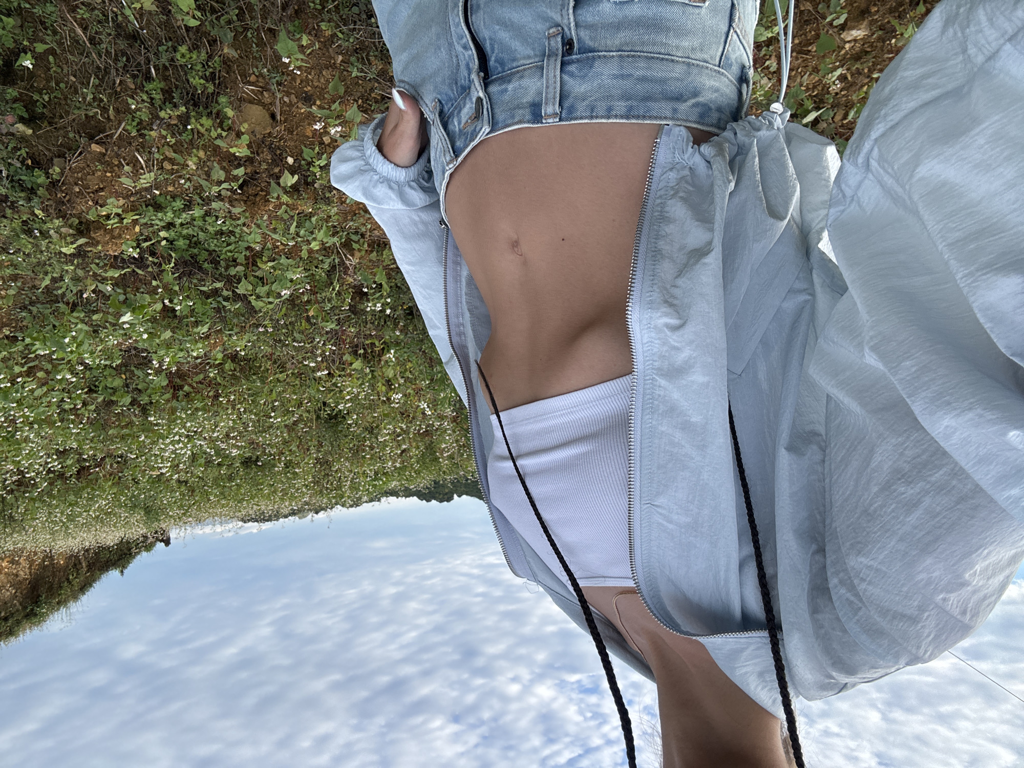 A woman stands in a field of blooming white flowers in Lũng Cú Commune, Vietnam, against a backdrop of a cloudy sky. She is wearing denim jeans and a light blue nylon jacket over a white tank top. The jacket is partially unzipped, revealing her midriff.