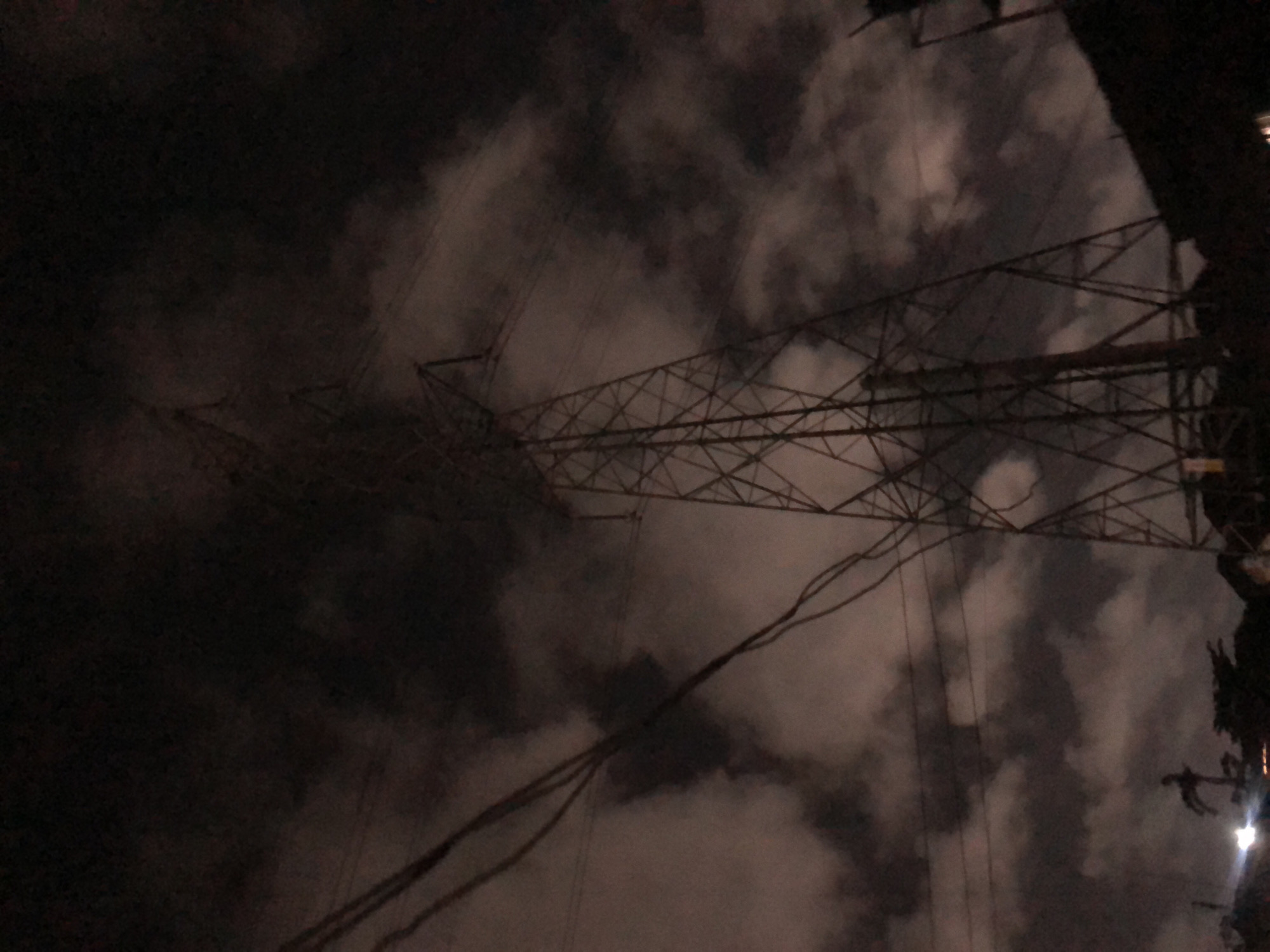 The image depicts a large electrical tower silhouetted against a dark, cloudy sky at night. Power lines are visible, extending from the foreground towards the tower. In the lower portion of the frame, a dimly lit landscape includes trees and possibly some structures. The overall scene suggests an industrial or semi-urban environment in Ado, Nigeria, at night, characterized by overcast skies and artificial lighting in the distance.