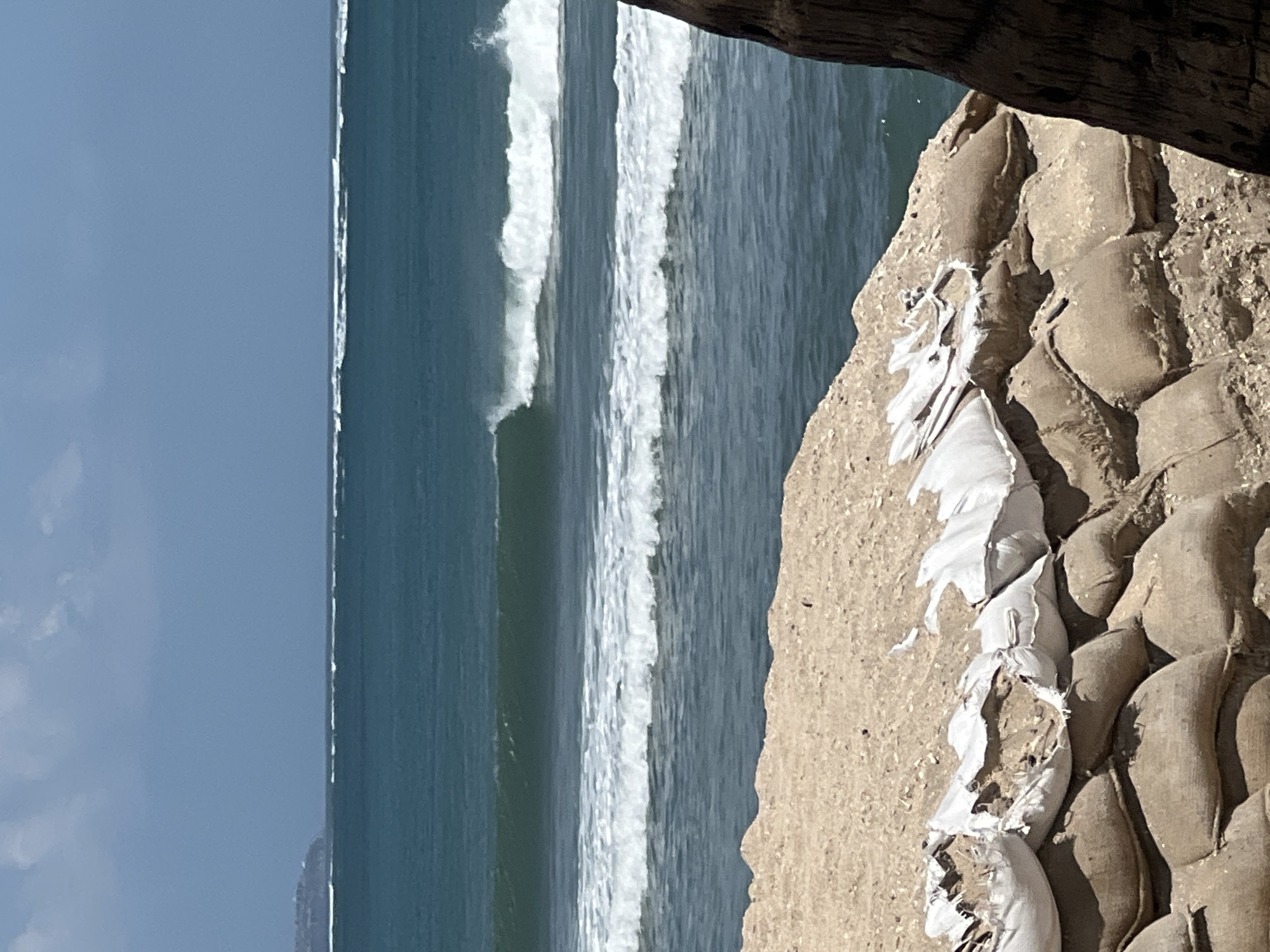 The image depicts a coastal scene with a focus on a barrier made of sandbags in the foreground. The sandbags are stacked in multiple layers, with some on top appearing to be white fabric bags filled with sand and debris, and those below being made of rougher, brown material. Behind the sandbags, the ocean is visible with several waves breaking. The water is a deep teal color, and the sky is a pale blue with some clouds. In the far distance, a faint outline of land with some structures can be seen on the horizon. The image does not contain any people or explicit actions, but the presence of the sandbags suggests a potential need for coastal protection, possibly due to erosion or flooding. The scene is consistent with a beach environment in Kuta, Indonesia, where such protective measures might be employed to safeguard the coastline.