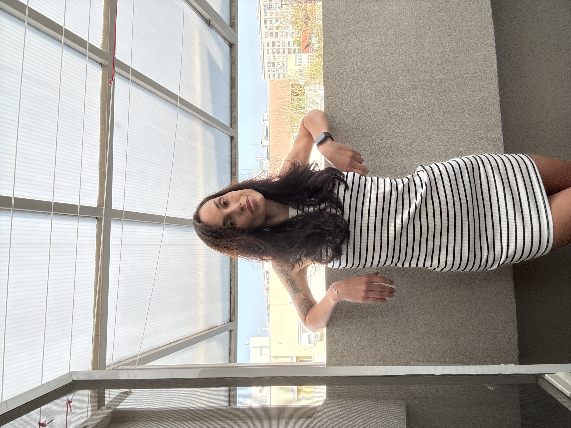 A young woman with long, dark hair is captured in a full-body shot, standing and leaning against a textured, light-colored wall or railing. Her body is slightly angled, and she gazes directly at the camera with a subtle smile. She wears a form-fitting, sleeveless dress featuring vertical black and white stripes. On her left wrist, she has a dark smartwatch, and on her right wrist, a delicate silver-toned bracelet, with a visible tattoo on her right forearm.
To her left, a structural element composed of metal frames holds large, translucent, ribbed panels, suggesting a windowed enclosure or roof, possibly part of a balcony or terrace structure. A thin red cord is visible near the top of these panels. To her right and in the background, a clear blue sky extends over an urban landscape, revealing numerous multi-story buildings and some green trees. The scene is brightly lit by natural daylight. No visible text is present in the image. The setting appears to be an urban environment, consistent with Pleven, Bulgaria.