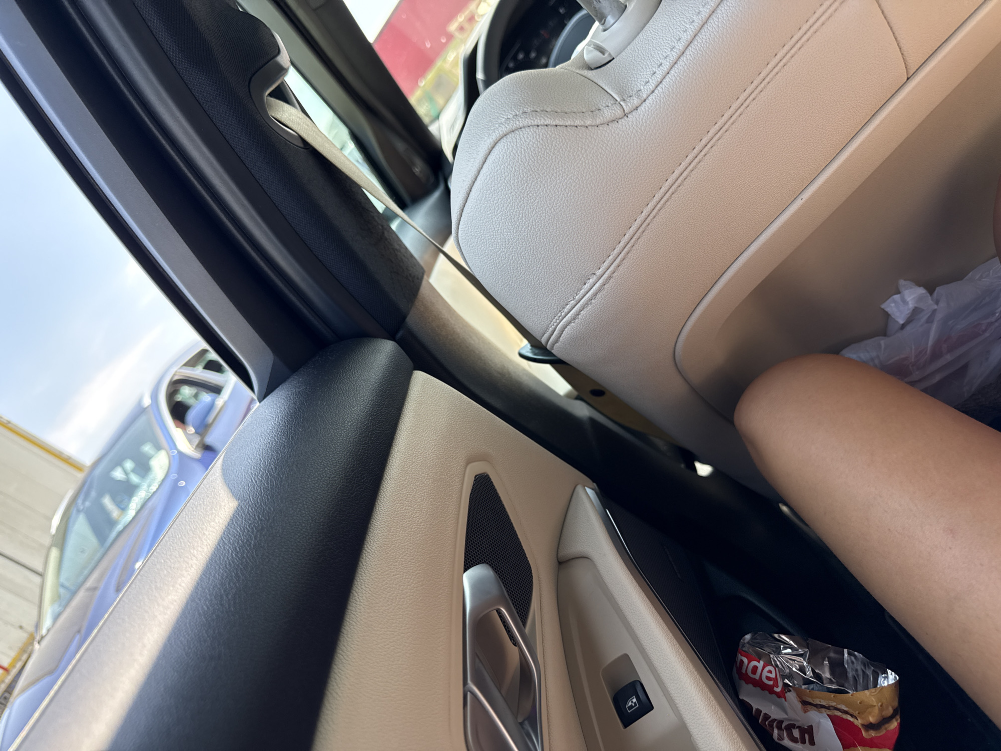 In Sanpetru, Romania, this image captures a close-up view from inside a car. The interior features light-colored leather seats and door panels. A person's leg is visible in the foreground, resting in the passenger seat. A partially unwrapped snack, possibly a biscuit or wafer bar, lies in a compartment to the left of the leg. Through the car's windows, a blue vehicle is seen parked outdoors, under a bright, sunny sky. The scene appears to be in a parking lot or on a street.