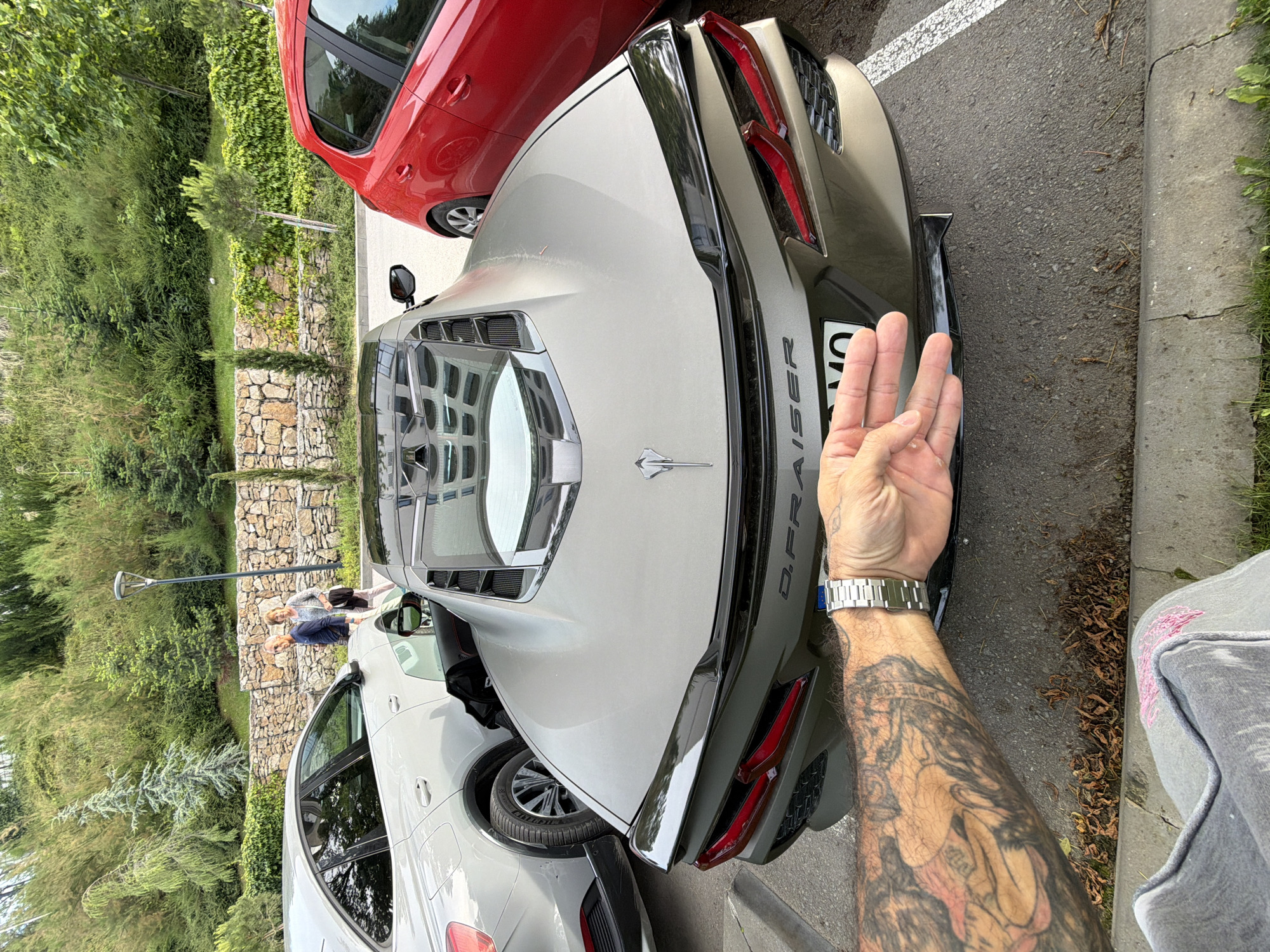 A person's tattooed arm and wrist wearing a silver watch is in the foreground, obscuring the license plate of a silver Corvette parked in a lot. The car's rear is visible, featuring distinctive taillights and a raised spoiler. A red car is parked to the right of the Corvette, and a white SUV is parked to the left. In the background, two people are walking near a stone wall and a street lamp, with lush green trees and vegetation covering a sloped hill. The location is identified as the city of Varna, Bulgaria.