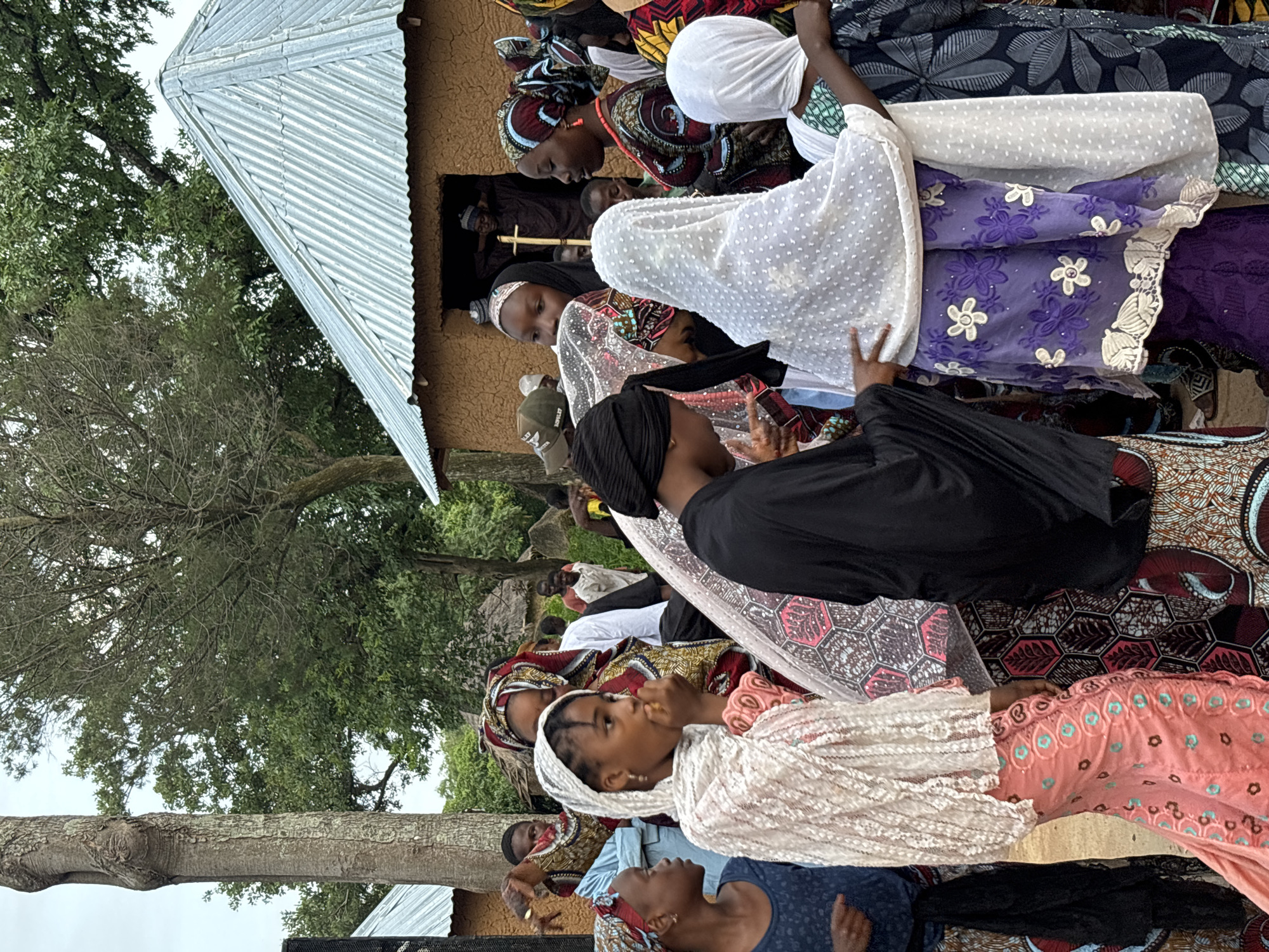 The image depicts a gathering of people, predominantly women and girls dressed in colorful traditional attire, outside of a mud-brick building with a corrugated metal roof. The scene appears to be in a rural or semi-rural setting, possibly a village in the city of Jos, Nigeria, given the architectural style and the clothing. The lighting suggests it is daytime, with overcast or cloudy skies.
In the foreground, a young girl in a white lace headscarf is eating something yellow. To her left, another young girl wears a blue shirt and a patterned headscarf. To the right of the first girl, a woman in a black headscarf is visible, and behind her, another woman wears a white dotted headscarf over a purple embroidered garment. Further back, more individuals are seen, some near the entrance of the building, where at least two men are visible. Trees and foliage are present in the background, adding to the outdoor setting. The overall impression is of a communal event or gathering.