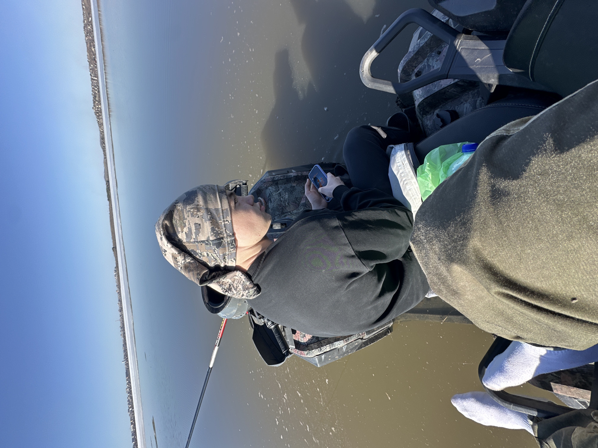 A person wearing a camouflage hat and dark clothing is seated on an all-terrain vehicle (ATV) near water, possibly a lake or wetland. They are holding a mobile phone and appear to be using it. A fishing rod is visible beside the ATV. Another person, partly visible, is seated beside them on the ATV. The sky is clear and blue. The location is unknown.