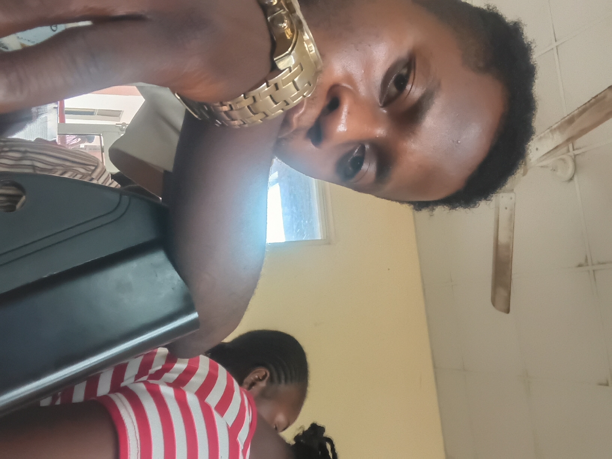 The image shows two people in what appears to be an indoor setting, possibly a room in Wukari, Nigeria. The orientation of the image is rotated.
In the foreground, a person's face is prominently displayed, with part of their arm visible. They are wearing a gold-colored watch on their wrist. Their skin tone is dark, and they have dark, curly hair. Their eyes are open and looking towards the viewer.
Below and slightly to the right of this person, another individual is partially visible. This person is wearing a red and white striped shirt. Their head is bowed, and their hair is styled in braids.
Behind the people, there are various objects and elements of the scene. A dark, possibly black, rectangular object is positioned to the left, obscuring part of the lower half of the first person. To the right, a window with light coming through is visible, suggesting it might be daytime. The walls of the room are a pale yellow or cream color, and a ceiling fan with light-colored blades is mounted on the ceiling. The overall impression is of a casual, everyday environment. No visible text is present in the image.