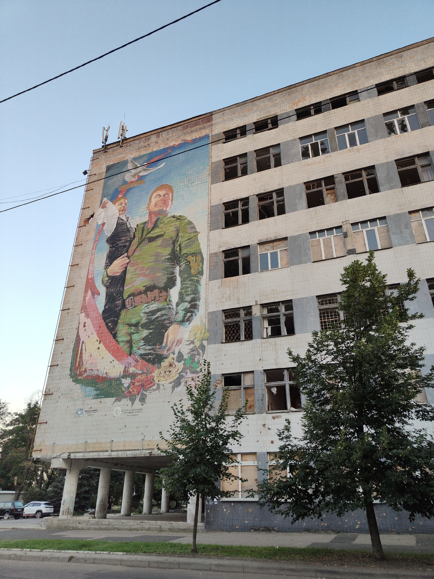 The image shows a large mural on the side of a multi-story, tiled building in Donetsk, Ukraine. The mural depicts a man in a military-style uniform holding a child, with a dove flying overhead and flowers at the bottom. The building has numerous broken windows and appears to be in a state of disrepair. The mural itself also shows signs of damage, with bullet holes visible. In the foreground, there is a street with a sidewalk and some trees. The sky is clear and blue.