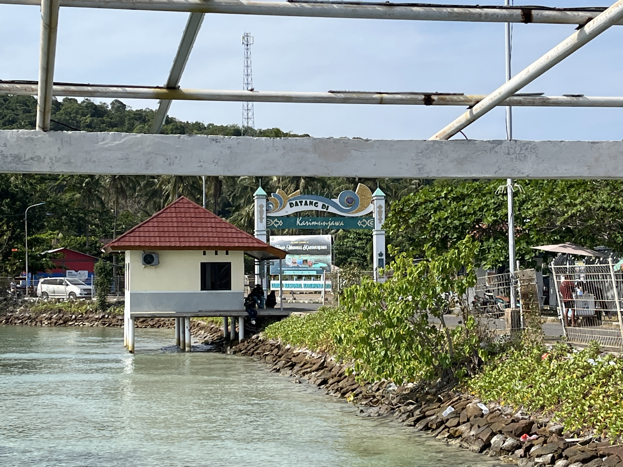 The image shows a waterfront scene in Karimunjawa, Indonesia, with a small, elevated building on stilts over the water. The building has a red tiled roof and a light-colored exterior with a dark window. A white minivan is parked on the shore behind a low stone wall. In the background, a large sign reads "DATANG DI KARIMUNJAWA" (Welcome to Karimunjawa) with decorative elements. A telecommunications tower is visible in the distance on a tree-covered hill. The waterfront is lined with rocks and lush green vegetation. Several people are seated on the wooden platform near the building. The water appears calm and light blue. Further down the shore, there are fences, parked motorcycles, a vendor stall with an umbrella, and more dense greenery. The sky is clear and blue.