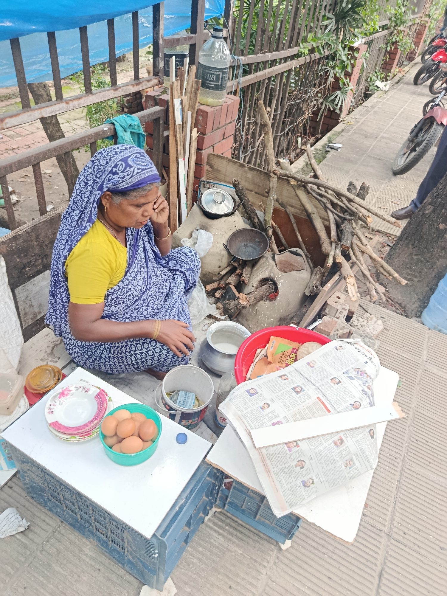 An elderly woman is seated on the pavement in Dhaka, Bangladesh, possibly preparing food. She wears a yellow blouse and blue patterned sari and sits beside a makeshift stove made of mud and bricks, with firewood stacked nearby. Cooking pots and pans are visible on and around the stove. A white crate topped with a white surface functions as a table, holding plates and a green bowl containing several brown eggs. Newspapers and other items are scattered on another crate beside her. A blue tarp is stretched above, and a metal railing separates the pavement from a lower area. Several parked motorcycles are in the background. A person is seen standing on the far right.