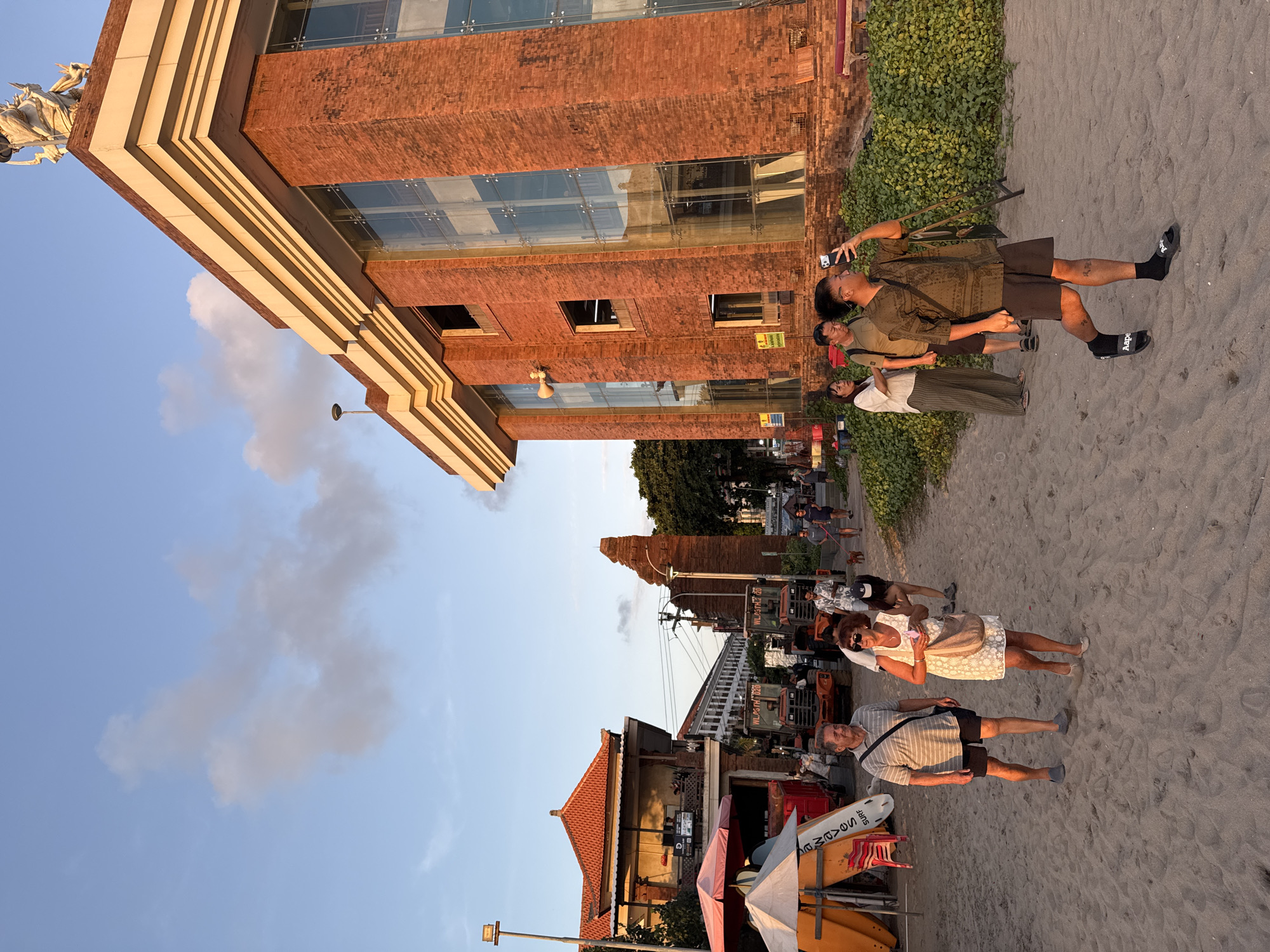 The image depicts a street scene in Kuta, Indonesia, at sunset. A large, modern brick building with glass windows dominates the right side of the frame, featuring stacked architectural layers and a statue of what appears to be a mythological figure on its roof. To the left, a traditional building with an orange tiled roof is visible, along with several orange tractors parked on a sandy surface that appears to be a beach or a roadside area. A group of people are walking on the sand, some appearing to be tourists. The sky is a clear blue with wispy clouds, and the setting sun casts a warm, golden light on the scene. Several surfboards are leaning against a structure on the left, suggesting proximity to the ocean.