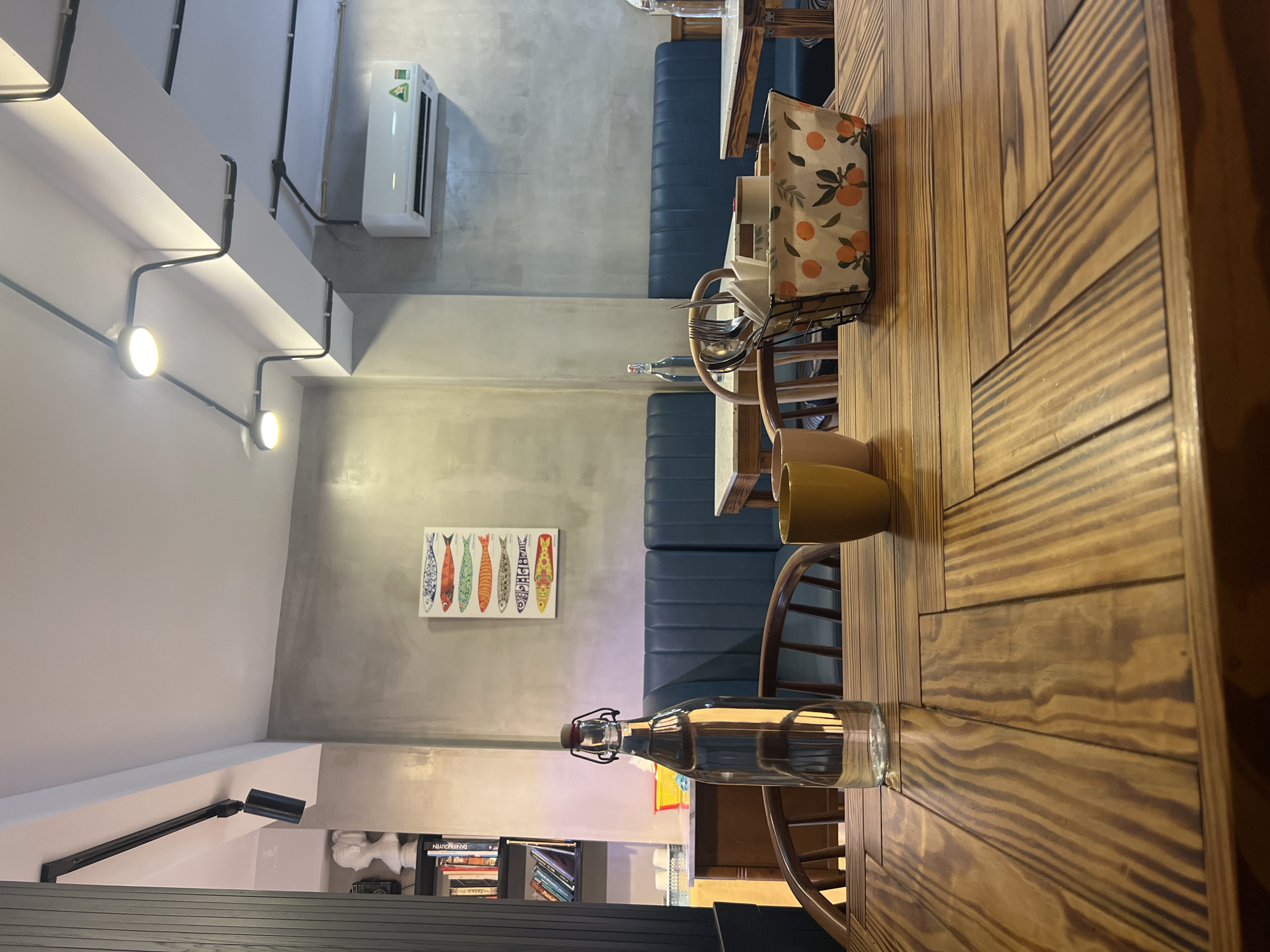 This image captures the interior of a modern cafe or restaurant in Bắc Nha Trang, Vietnam, taken from a low angle across a wooden table. No people are visible, suggesting a quiet moment.
The scene is dominated by a richly textured wooden table, featuring planks arranged in a decorative pattern. On the table, a clear glass bottle filled with water, two ceramic mugs (one yellow, one brown), and a metal caddy holding cutlery wrapped in a fabric napkin with an orange fruit print are neatly arranged.
The background reveals a minimalist yet inviting space. The walls are a cool, concrete-like grey, contrasting with a clean white ceiling and a prominent white beam from which exposed black conduit lighting fixtures extend, casting warm light. An air conditioning unit is mounted high on the far wall. Along the main wall, comfortable blue upholstered bench seating is arranged. A vibrant rectangular painting depicting six colorful, patterned fish adds a pop of color and artistic flair to the grey wall. Below it, a dark bookshelf houses various books, including one with "DAN NGUYEN" visible on its spine, and decorative items like a white bust and a vintage camera. In the distant background, hints of a service area with white crockery can be seen.
The atmosphere appears calm and contemporary, with a blend of industrial and rustic elements, suitable for a relaxed dining experience.