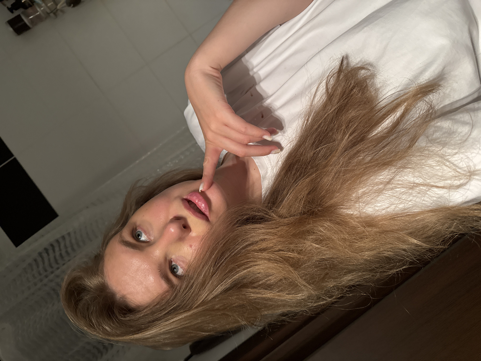 A young woman with long, light brown hair, looking upwards with a thoughtful expression, is captured in a medium shot. Her right index finger is placed near her lips, and her left hand rests on her chest. She is wearing a white t-shirt. The background appears to be a bathroom, with a shower curtain visible on the left and tiled walls. The lighting is dim, suggesting an indoor setting, possibly a residential bathroom. The location is not specified in the image.