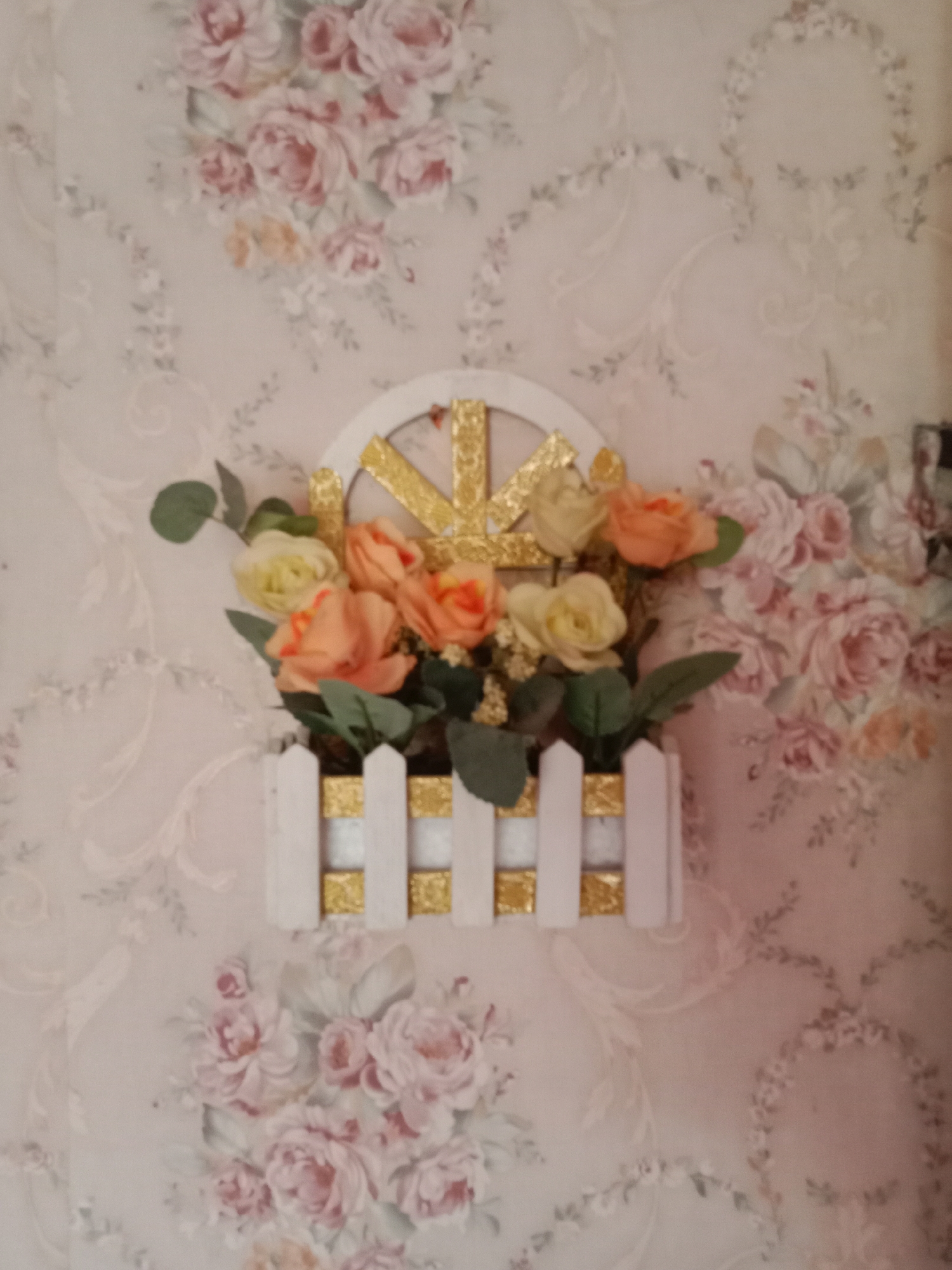A wall adorned with floral wallpaper serves as the backdrop for a decorative hanging. The wallpaper features a pattern of soft pink and white roses, interspersed with delicate green foliage. Mounted on the wall is a white, arched decorative element with gold accents. Nestled within this frame is an arrangement of artificial flowers, primarily in shades of peach and pale yellow, with some green leaves. The flowers are contained in a white box that resembles a picket fence, also decorated with gold squares. The scene depicts an indoor setting, likely a home or a business, and is not associated with any specific outdoor location within Kutosari, Indonesia, as no geographical markers are present.