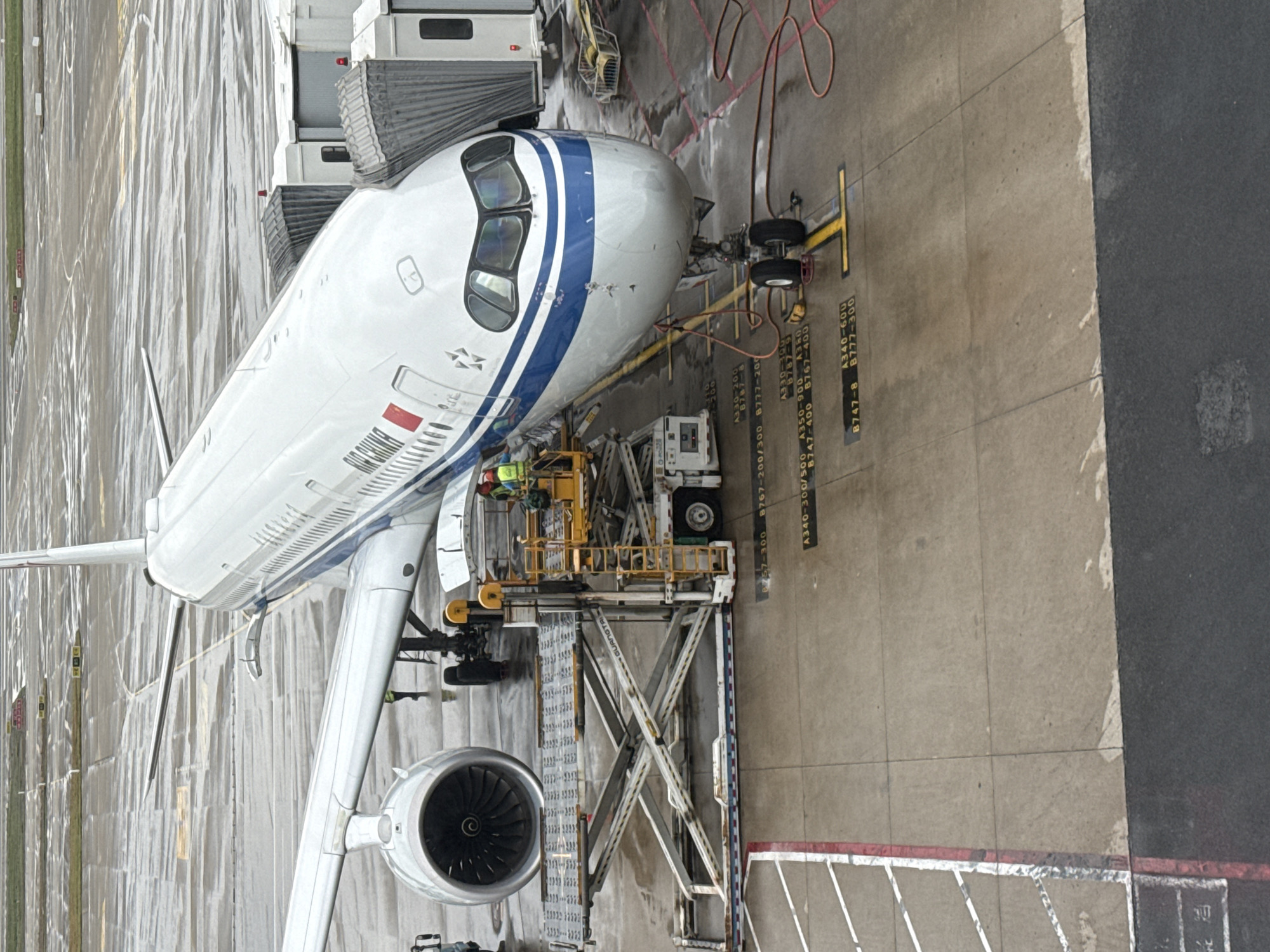 In Pudong, China, an Air China airplane is parked at a gate, connected to a jet bridge. The tarmac is wet, indicating recent rain. A baggage loader is positioned near the aircraft's cargo hold, with two workers in high-visibility vests attending to it. The plane's engines are visible, and the cockpit windows show reflections. Various ground markings on the tarmac indicate aircraft parking positions. The scene appears to be a typical airport operational environment.