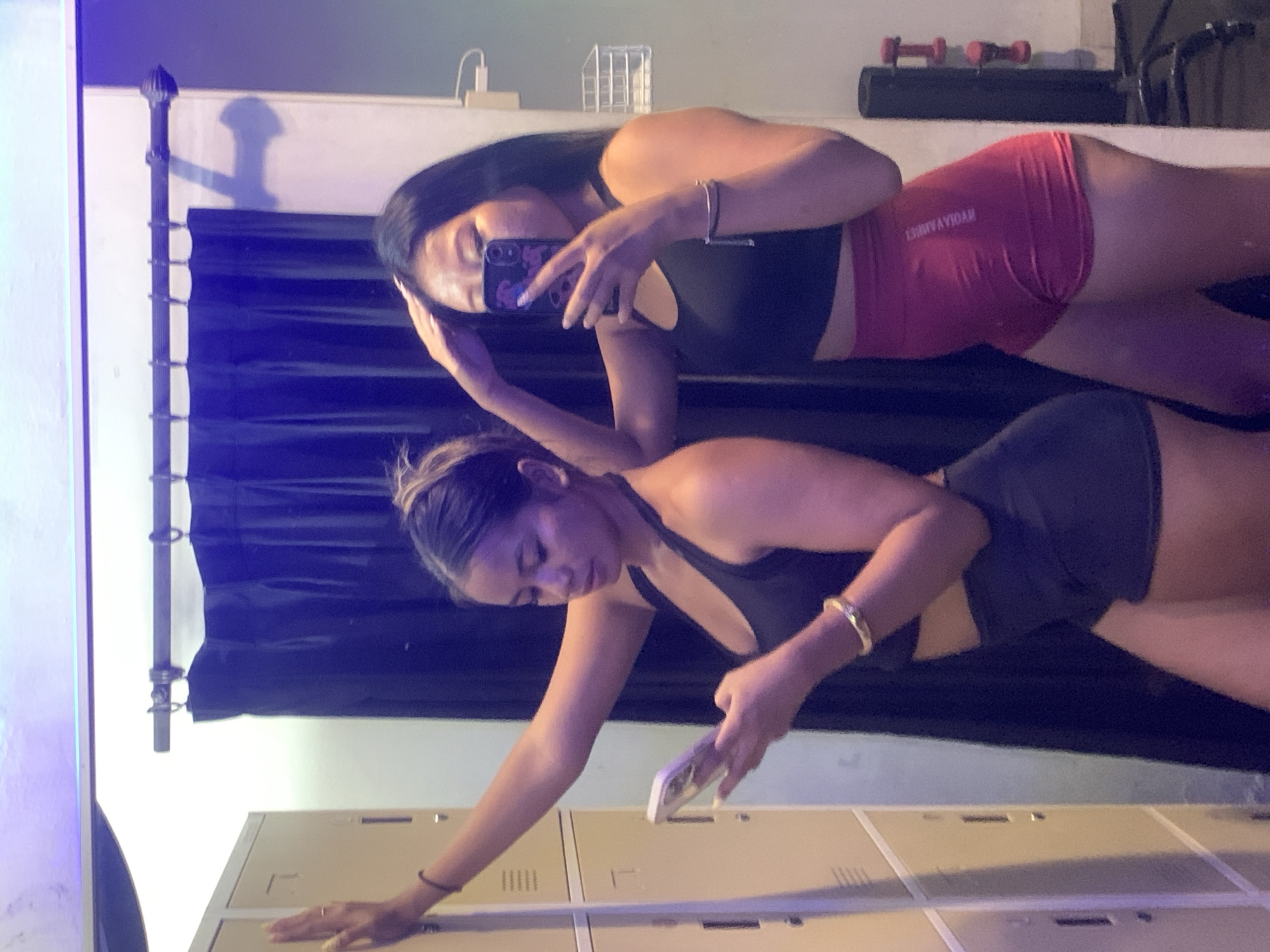 Two young women are taking a mirror selfie in what appears to be a locker room. The woman on the left is wearing a black sports bra and black shorts, and is holding a white phone. The woman on the right is wearing a black sports bra and red shorts, and is holding her phone to take the picture. She has her other hand on her head. Behind them, there are lockers and a dark blue curtain. The lighting is dim and appears to have a blueish hue. It is not possible to determine the city from the image itself, however, it is stated that the image is located in Denpasar, Indonesia.