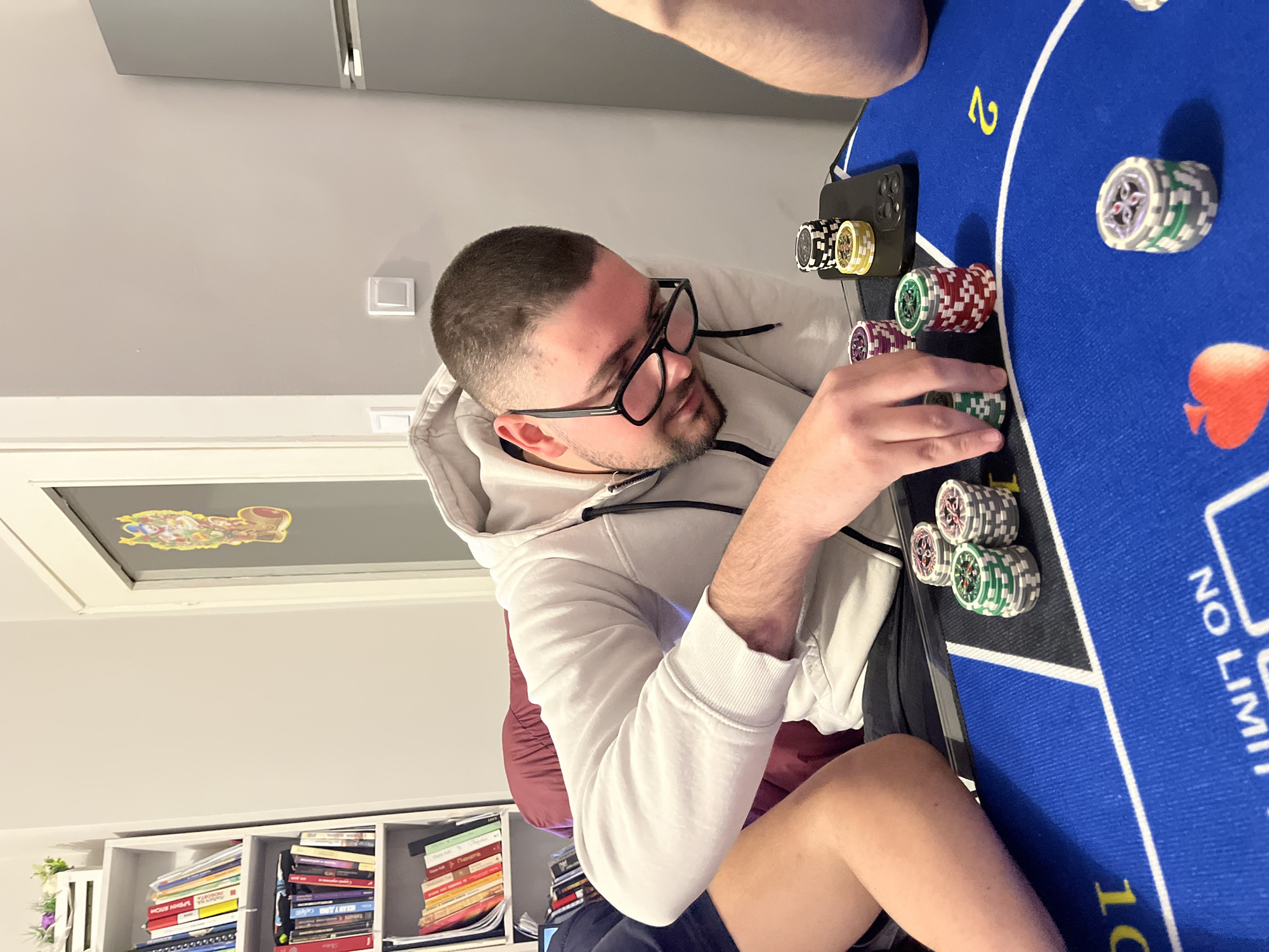 A man with a shaved head, a beard, and glasses is sitting at a blue poker table, stacking poker chips. He is wearing a white hoodie and dark shorts. There are several stacks of poker chips on the table, along with a black phone. The table has "NO LIMIT" printed on it. In the background, there is a white bookshelf filled with books, a light-colored wall, and a silver refrigerator. The location appears to be indoors, potentially in an apartment or house. I am unable to confirm if the image was taken in Burgas, Bulgaria, as there is no geographical information available in the image.