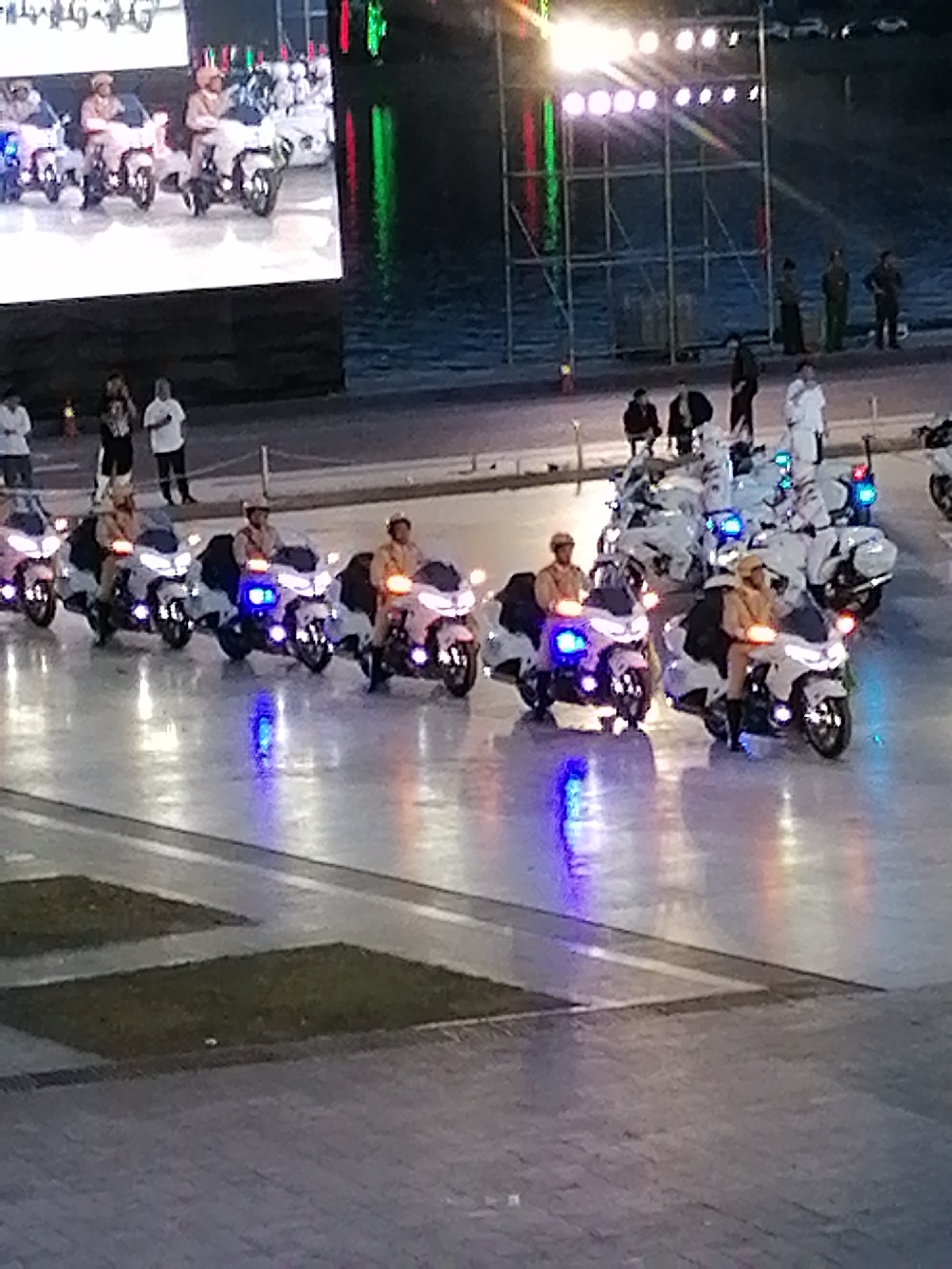 This image shows a procession of police officers on motorcycles, likely participating in a parade or ceremonial event. The motorcycles are white with blue and red lights. The officers are wearing light-colored uniforms and helmets. The scene appears to be at night, with the motorcycles' lights reflecting on a wet surface, possibly a street or plaza. In the background, there are large screens displaying images of people on motorcycles, and a structure with lights. There are also spectators visible in the background. The location is identified as the city of Da Lat, Vietnam.