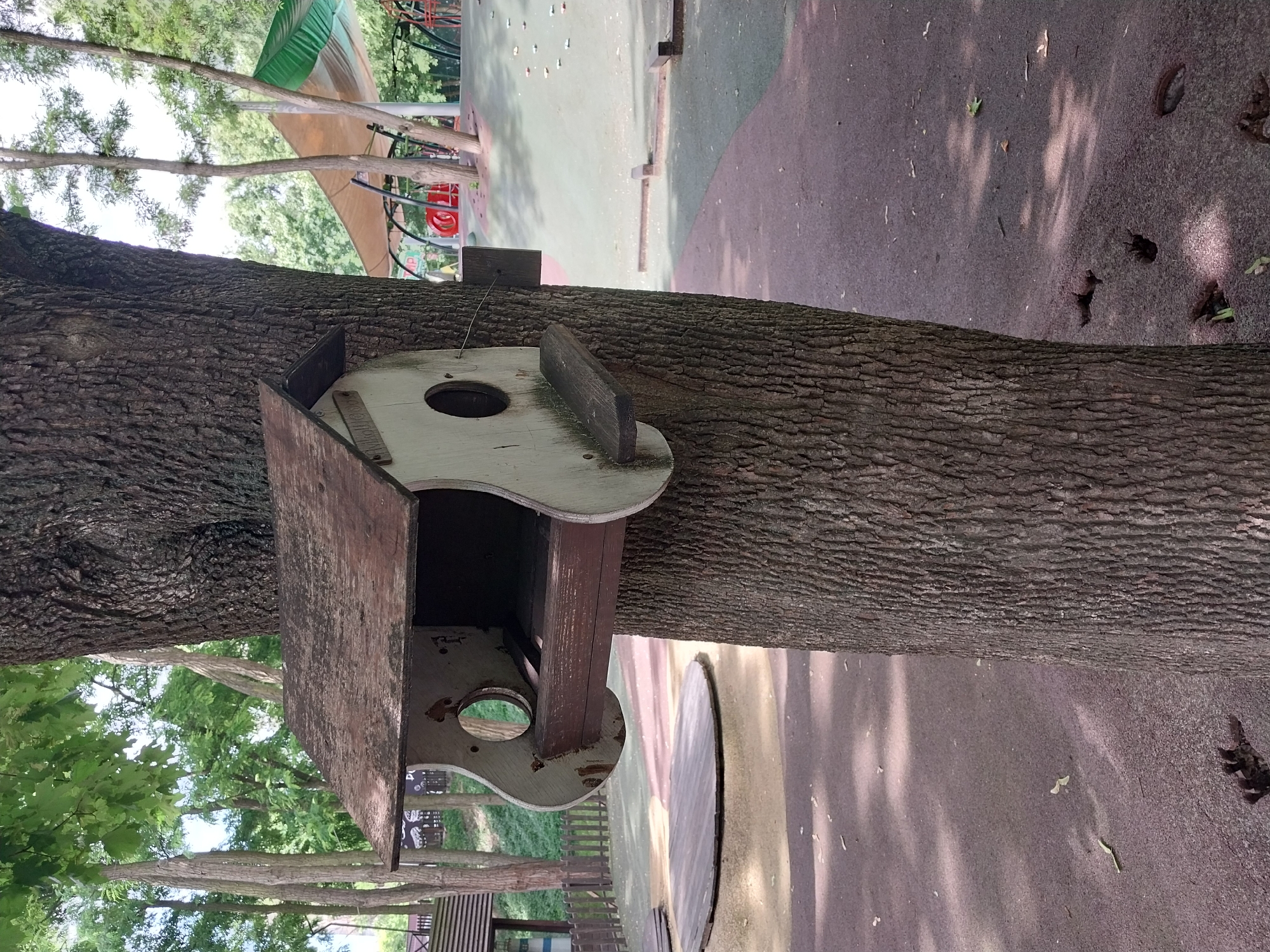A weathered, light-colored wooden birdhouse with a dark brown roof is mounted on the trunk of a large tree. The birdhouse has two circular openings, one on the left with a reflective surface, and a shelf-like perch extending from the bottom. The tree is rough-barked and centrally located in the frame. In the background, a playground area is visible, featuring a green and beige canopy structure, red play equipment, and a sloped surface with various play features. The ground in the foreground is a dark, textured surface with shadows and scattered leaves. The scene appears to be in an outdoor public space.