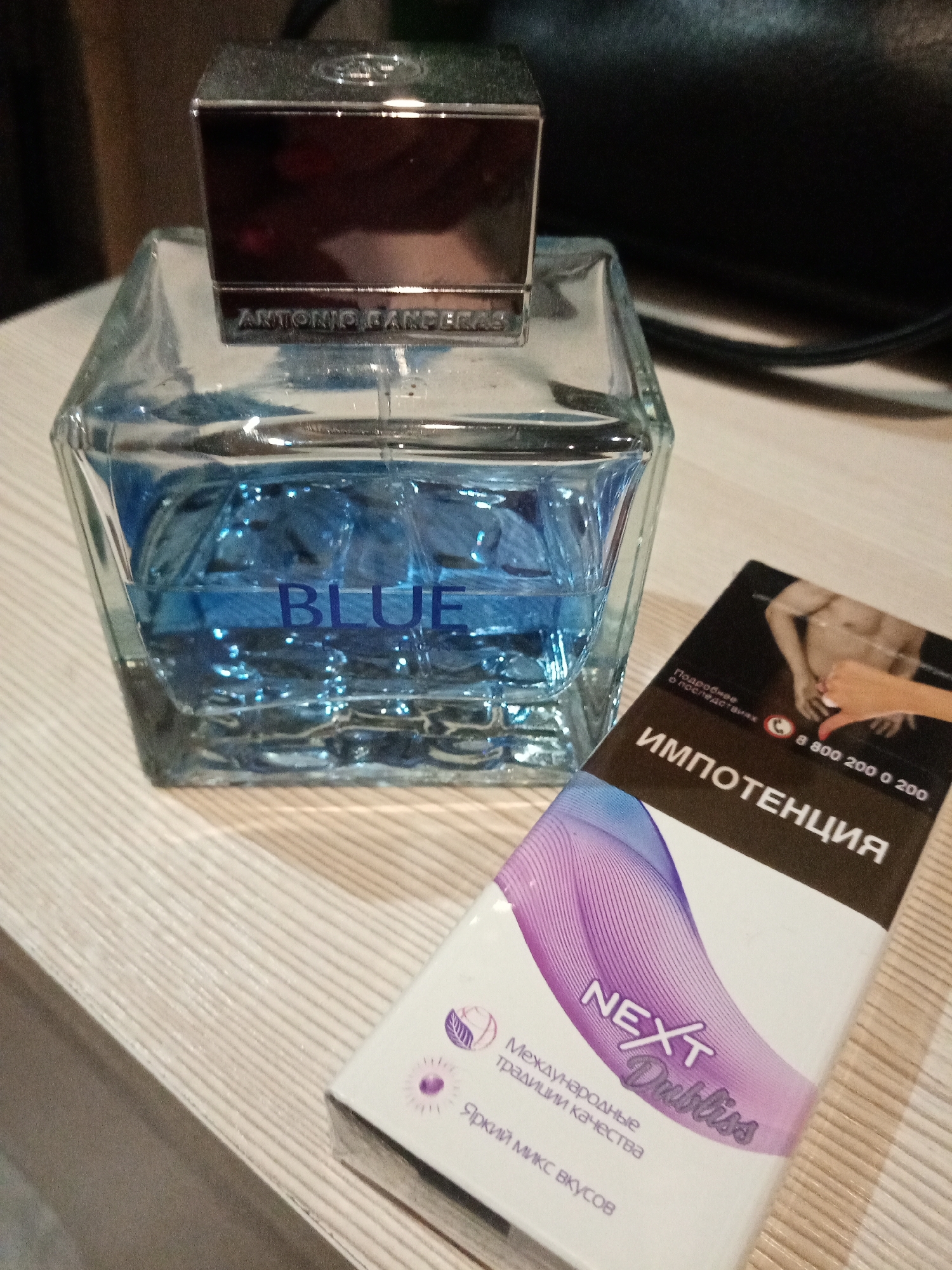 The image shows a bottle of "Blue" by Antonio Banderas perfume next to a pack of Next Dubliss cigarettes. The cigarette pack is white with purple accents and features a graphic image related to impotence. The table appears to be a light wood grain surface. The background is dark, with a black bag or object partially visible. The image was captured in Elektrostal, Russia, as the text on the cigarette pack is in Russian. There are no people visible in the image.