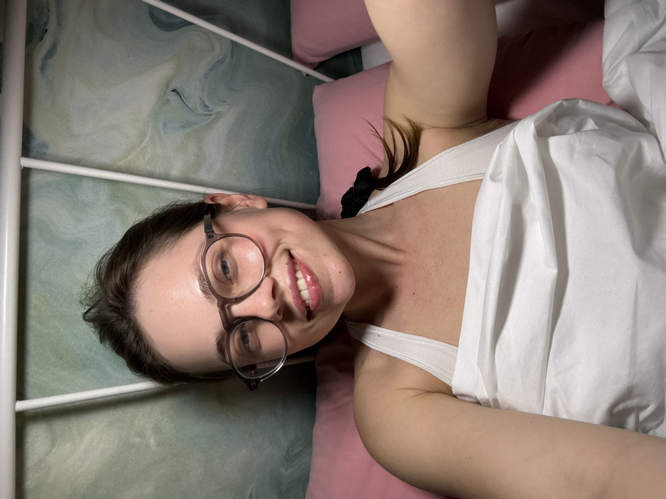 The scene, located in Scafati, Italy, depicts a female individual lying in a bed. She is looking directly at the camera with a smiling expression, displaying visible dimples and a glossy finish on her lips. She has light brown hair, pulled back, and wears round-rimmed glasses with a translucent frame. Her attire consists of a white halter-neck top, and a white sheet or blanket is draped over her, covering her lower chest. She is supported by pink pillows. Behind her, a wall or headboard features an abstract swirling pattern in green and white tones, framed by a white metal bed structure. A black hair tie is visible on her left shoulder.