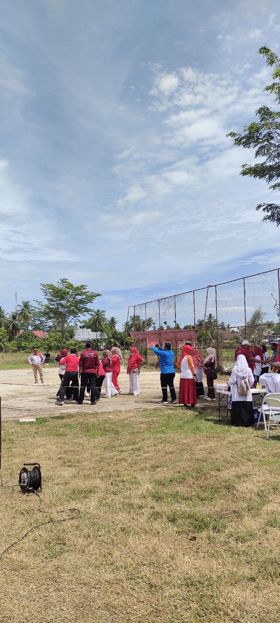 In the city of Meucat, Indonesia, a group of people in red and white attire are engaged in an outdoor activity. The scene appears to be a sports field with a fence in the background. Some individuals are observing, while others are actively participating in what seems to be a communal gathering or event. The sky is clear with scattered clouds, indicating fair weather. The ground is covered with dry grass, and trees are visible in the distance. A spool of cable is present on the left side of the frame, suggesting the presence of some equipment, possibly for sound or lighting. The overall atmosphere is casual and community-oriented.