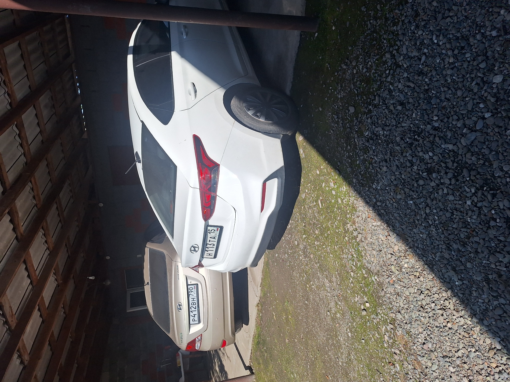 Two Hyundai cars are parked under a corrugated metal roof structure, possibly a carport or an open garage. The car in the foreground is white, and the car behind it is beige. Both vehicles have license plates. The beige car has a license plate that reads "P412BH 790 RUS". The white car has a license plate that reads "H113TA 15 RUS". The ground is covered with gravel and some patches of grass. The scene appears to be outdoors, in a location that could be residential. The presence of the license plates with Russian regional codes suggests the location is within Russia. Given the provided context that this image is from Chermen, Russia, it is safe to infer the scene is located in Chermen. The image depicts a static scene with no discernible activity or individuals.