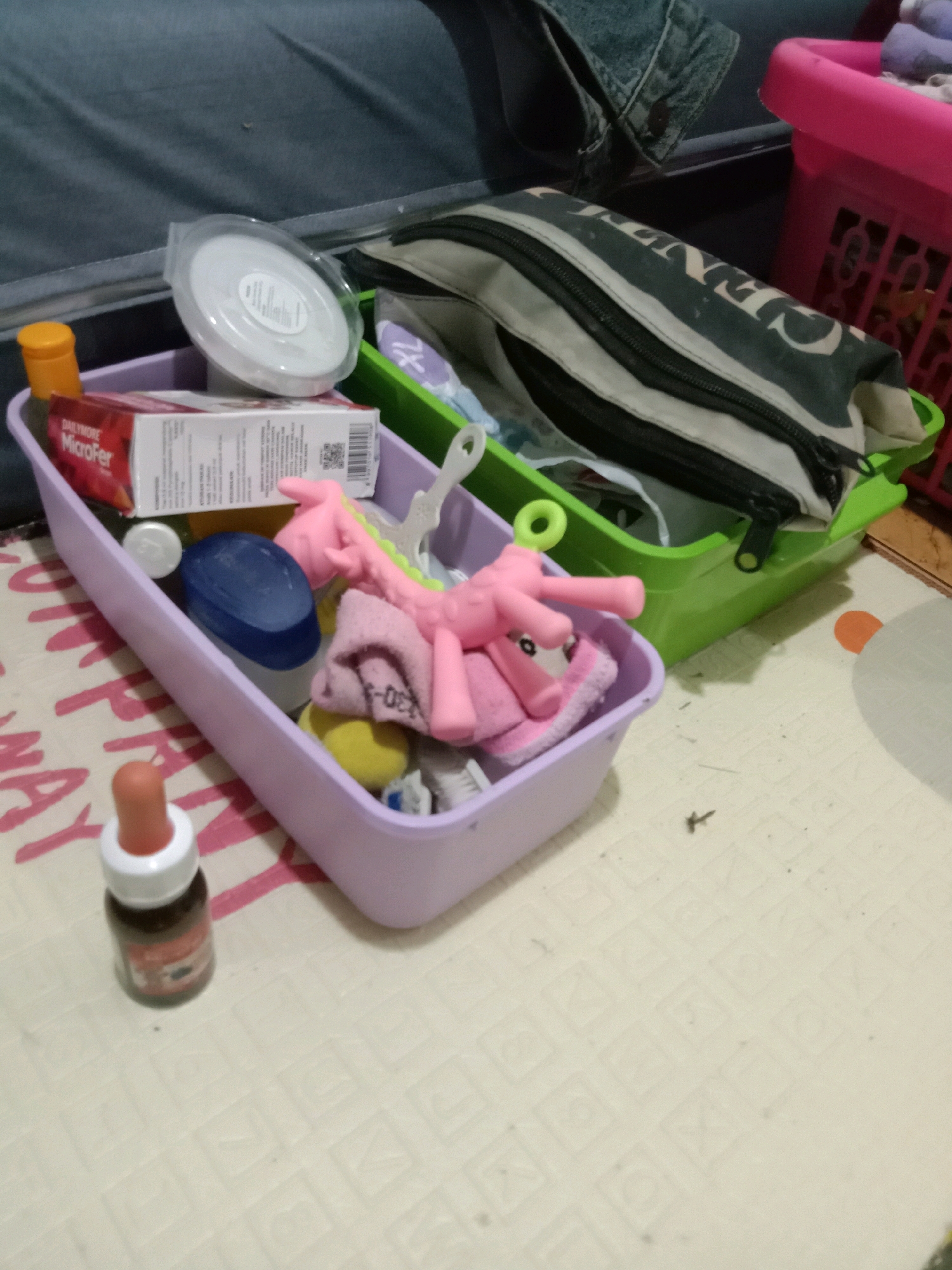 In the city of Jumput, Indonesia, a cluttered scene unfolds. A pale pink plastic container is filled with various items. Among them, a pink toy horse with a small pink cloth tucked underneath, a blue oval object, a white circular container with a lid, and a box labeled "DAILYMORE MicroFer" are visible. To the left of the container, an orange-capped bottle sits on a patterned mat. To the right and slightly behind, a green plastic container holds a zippered pouch, partially obscuring other items. In the background, a pink basket and a section of a dark blue fabric are visible. The overall impression is one of disarray within a domestic setting.