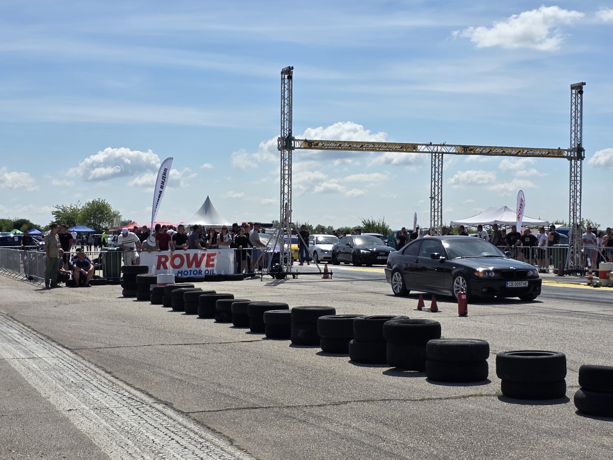 The image depicts a drag race event at the Akatsievo airfield, located in Akatsievo, Bulgaria. In the foreground, stacks of tires are arranged along the side of the asphalt track, serving as a safety barrier. A black BMW coupe, displaying the license plate CB 0097 HC, is positioned on the track, indicating its participation in the race. Several spectators are visible behind a metal fence, observing the event. White tents and banners, including one with the "ROWE MOTOR OIL" logo, are set up in the background, suggesting a commercial sponsorship of the event. The sky is clear with scattered clouds, indicating fair weather conditions for the outdoor activity. A large gantry structure with lights and equipment spans the track, likely marking the start or finish line of the race. The scene captures a moment of anticipation before or during a racing competition.