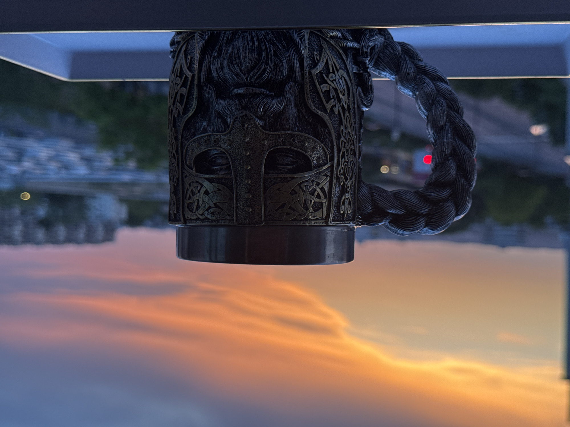 A close-up, eye-level shot captures a Viking-themed mug with a braided handle and intricate carvings on its body. The mug is positioned in the foreground on a light-colored tray, with a blurred city skyline visible in the background. The sky above displays a vibrant sunset with hues of orange, yellow, and blue, indicating an outdoor setting, possibly on a balcony or window ledge. The city lights twinkle in the distance, suggesting it is dusk or early evening. The overall scene is still and serene, with the dramatic sky providing a striking backdrop to the detailed mug.
