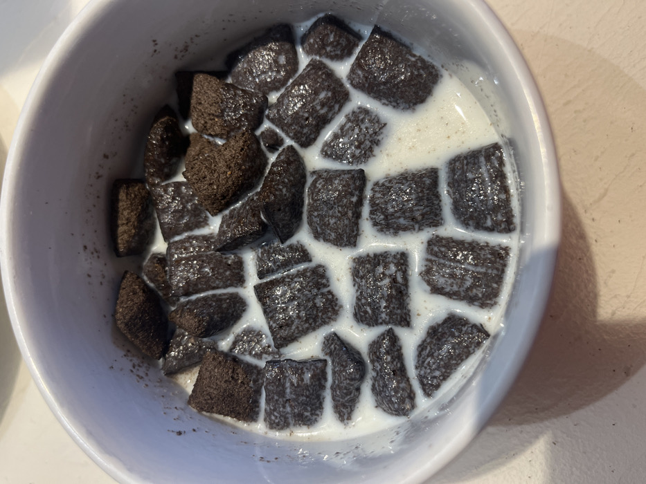 A white ceramic bowl is depicted containing a serving of dark brown, square-shaped cereal pieces submerged in white milk. The cereal pieces exhibit a somewhat porous texture, and some appear to be absorbing the milk, showing lighter areas. Small, dark crumbs are visible adhering to the interior rim of the bowl and dispersed within the milk. The milk partially covers the cereal, showing minor discoloration from interaction with the cereal. The bowl casts a shadow on a light-colored surface, likely a tabletop, beneath it. The scene is located in the city of Ancenis, France. No people or active interactions are visible.