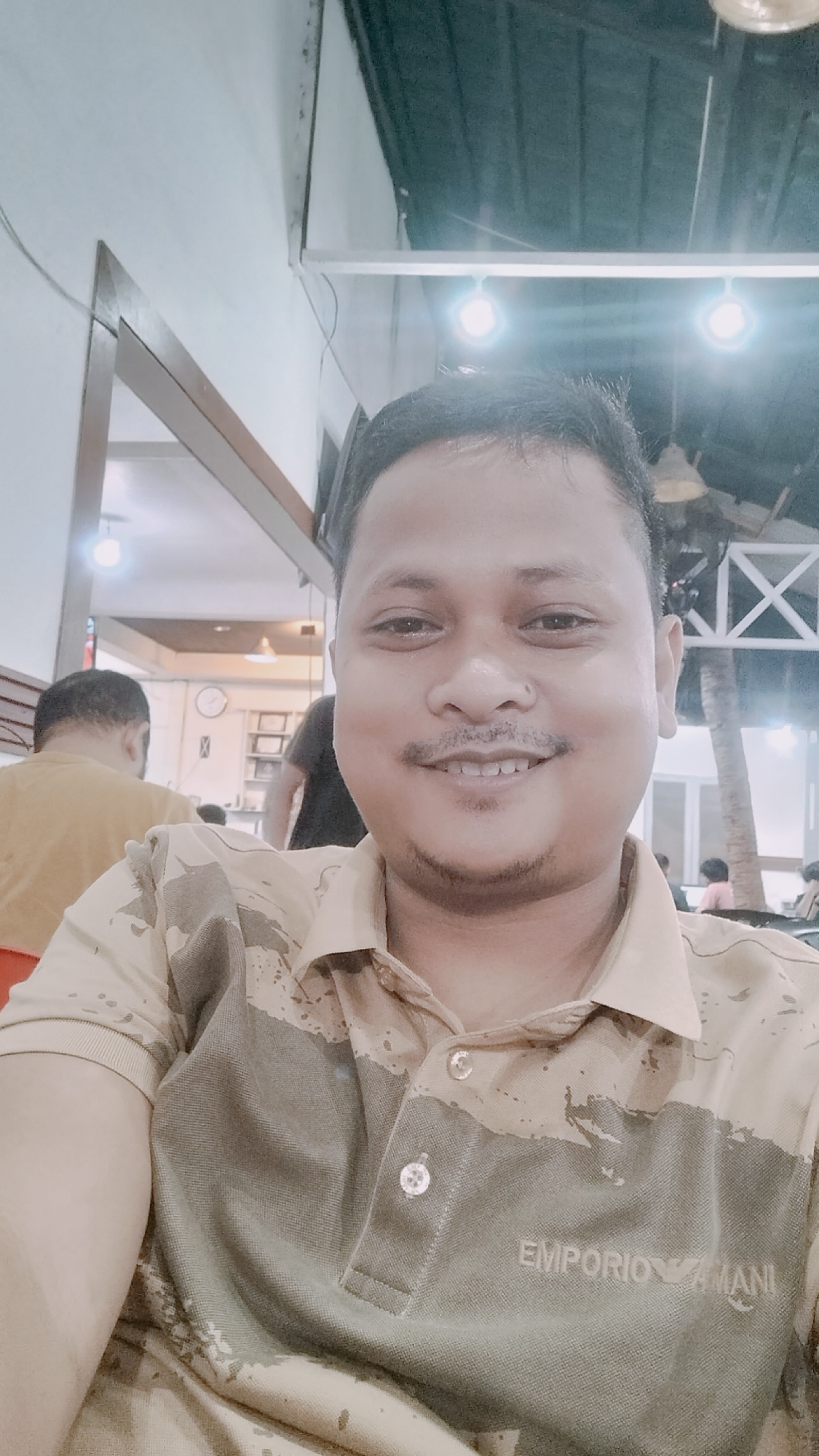 The image depicts an adult male taking a selfie in an indoor establishment, identified as being in Lhokseumawe, Indonesia. The subject, positioned centrally, is smiling directly at the camera. He has dark, short hair, a mustache, a short goatee, and a small stud piercing on his left nostril. He is wearing a two-toned polo shirt with a beige or light tan upper section and an olive green or dark tan lower section, separated by an abstract, mottled pattern. The brand "EMPORIO ARMANI" with its eagle logo is visible in white text on the lower right portion of the shirt.
The background reveals an indoor public space, likely a restaurant or cafe, with several other individuals and architectural features. To the subject's left, another man with short dark hair is visible from behind, wearing a mustard yellow t-shirt and appearing to be seated. Beyond him, a partial view of a white wall includes a circular clock with dark markings and a set of shelves with indistinct objects. To the subject's right, a distinctive white or light-colored wooden structure with an "X" pattern is prominent, suggesting a decorative partition or support beam. Behind this, the trunk of a palm tree is visible, indicating either an open-air section or a large window looking outdoors. Further back on the right, two additional blurred figures are seated, one wearing a pink/red garment. A man in a dark shirt is partially visible, walking away from the camera in the mid-background. The ceiling features visible conduit or wiring and multiple bright, unshaded artificial light sources, casting a cool-toned illumination throughout the space, with some areas appearing slightly overexposed.