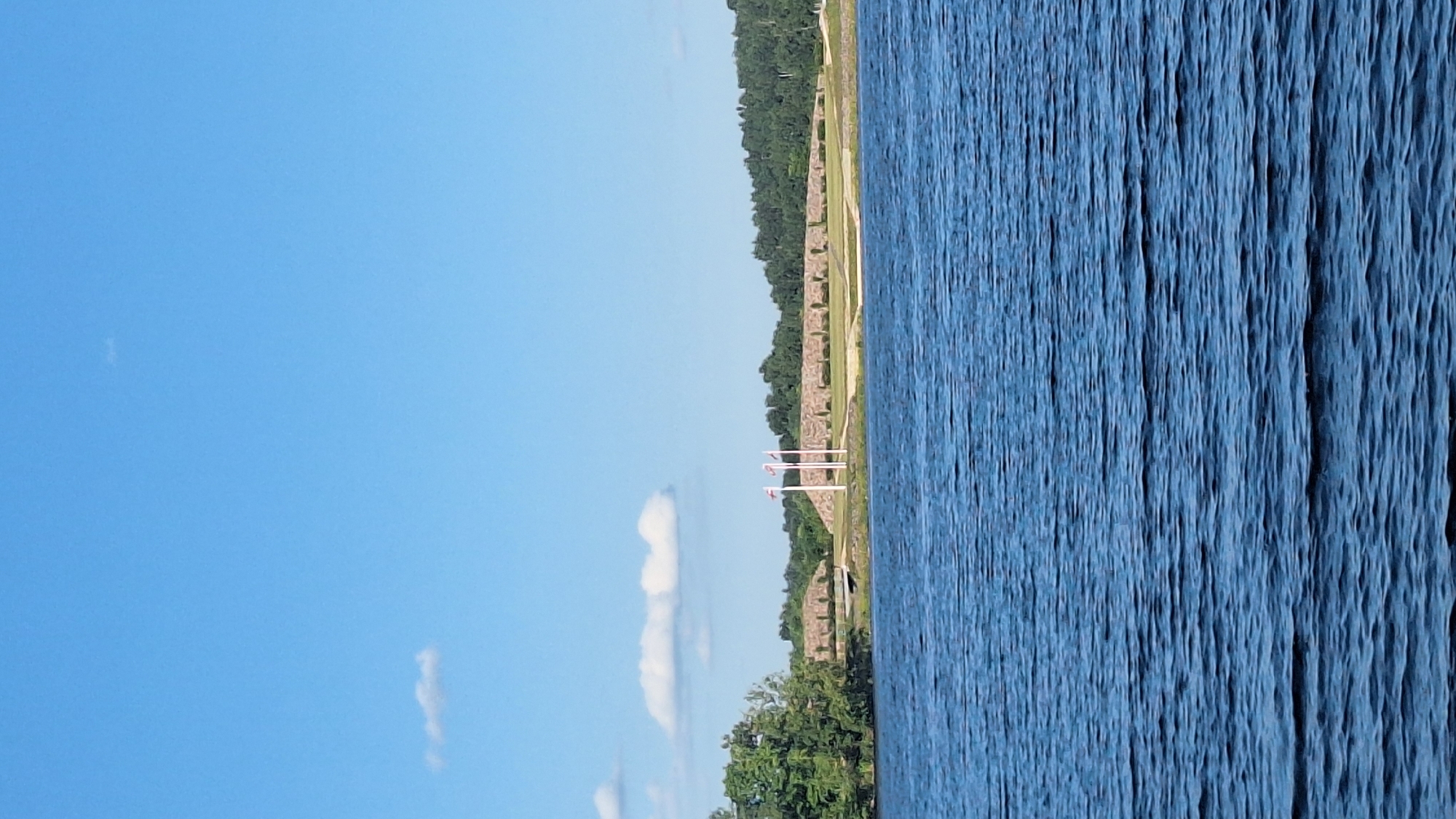 The image shows a wide body of rippling blue water in the foreground, stretching towards a distant shore. On the shore, a stone-built structure resembling a fortification or embankment dominates the mid-ground. Three flagpoles stand prominently in front of this structure, each flying a white flag with a red cross. Beyond the fortification, a dense line of dark green trees forms a backdrop, suggesting a natural landscape. The sky above is a clear, bright blue with a few wispy white clouds scattered across it. The overall scene appears to be a calm, outdoor environment, possibly a park or historical site near a body of water. The location is identified as the historic fort of Vecpiebalga in Sece, Latvia.
