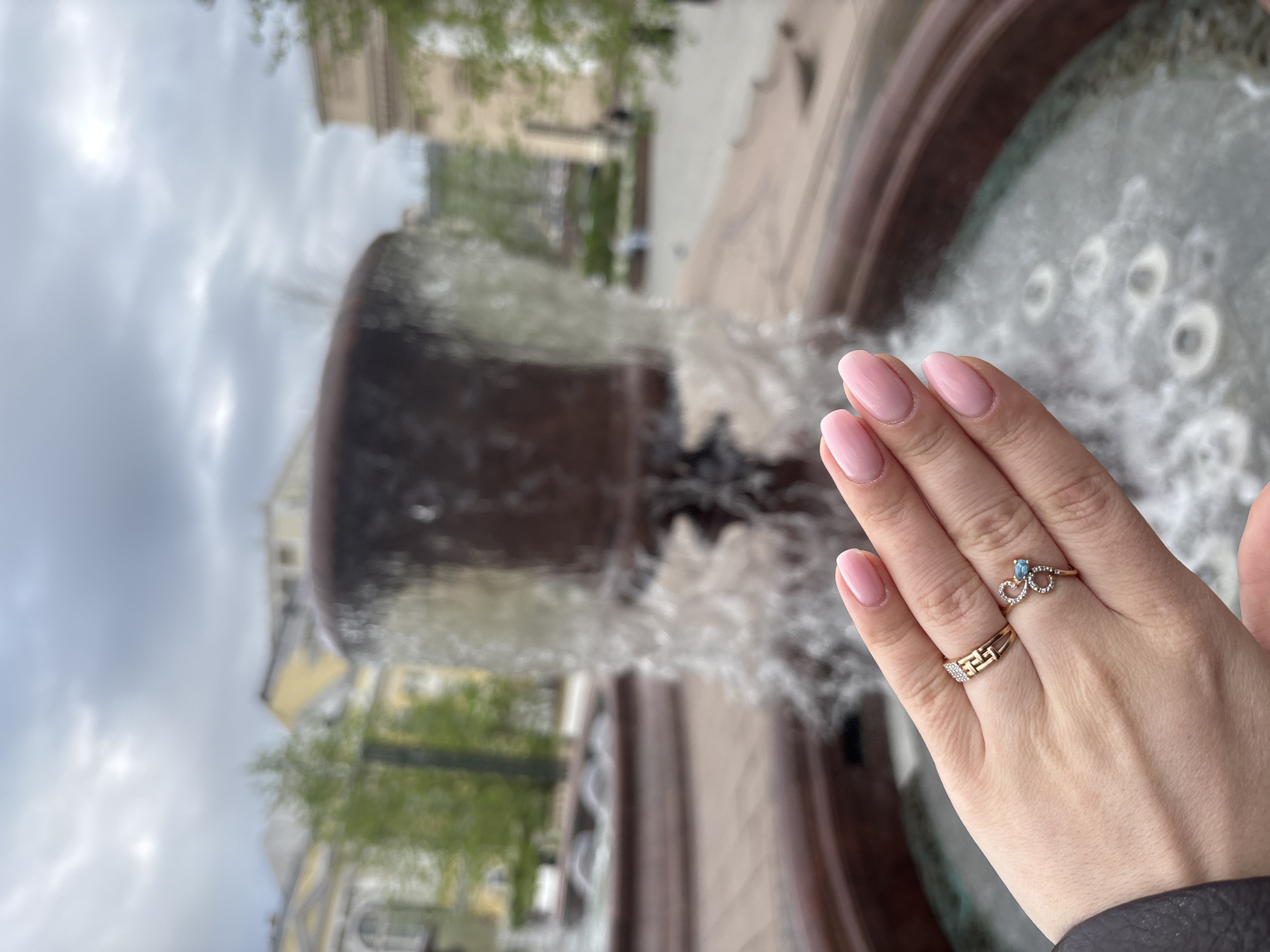 A right hand with two rings on the ring finger and the middle finger, held in front of a fountain. The nails are painted with a light pink polish. The ring on the ring finger is gold, with a blue gem, and a heart design with small stones. The ring on the middle finger is gold, with a textured design and small stones. The fountain in the background is large and made of a dark material, with water cascading down. The background includes buildings and trees, suggesting an outdoor, urban setting. The location is Moscow, Russia.