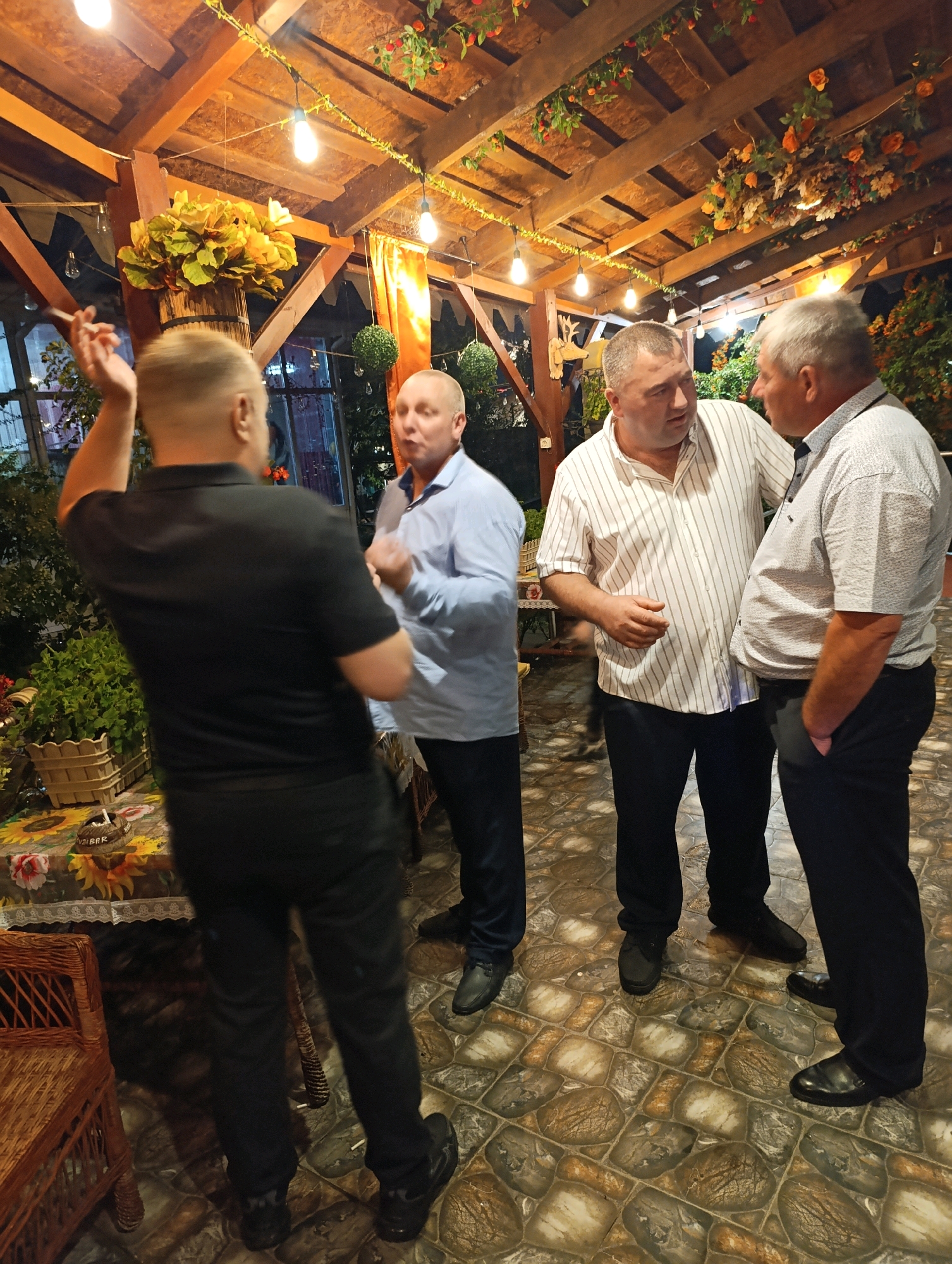 Four adult males are gathered in an outdoor covered patio area at night. The patio has a wooden ceiling with exposed beams and decorative lights strung across it, along with artificial floral garlands. The floor is paved with patterned tiles. One man, seen from behind and slightly to the left, is wearing a black polo shirt and black pants, holding a cigarette in his raised right hand. Another man, in the center left, is wearing a light blue collared shirt and dark pants. To his right, a man in a white horizontally striped collared shirt and dark pants is gesturing with his hands as he speaks to a fourth man, who is on the right, wearing a light gray patterned collared shirt and dark pants, with his left hand in his pocket. There are potted plants and decorative elements visible around the patio. The scene appears to be a social gathering.