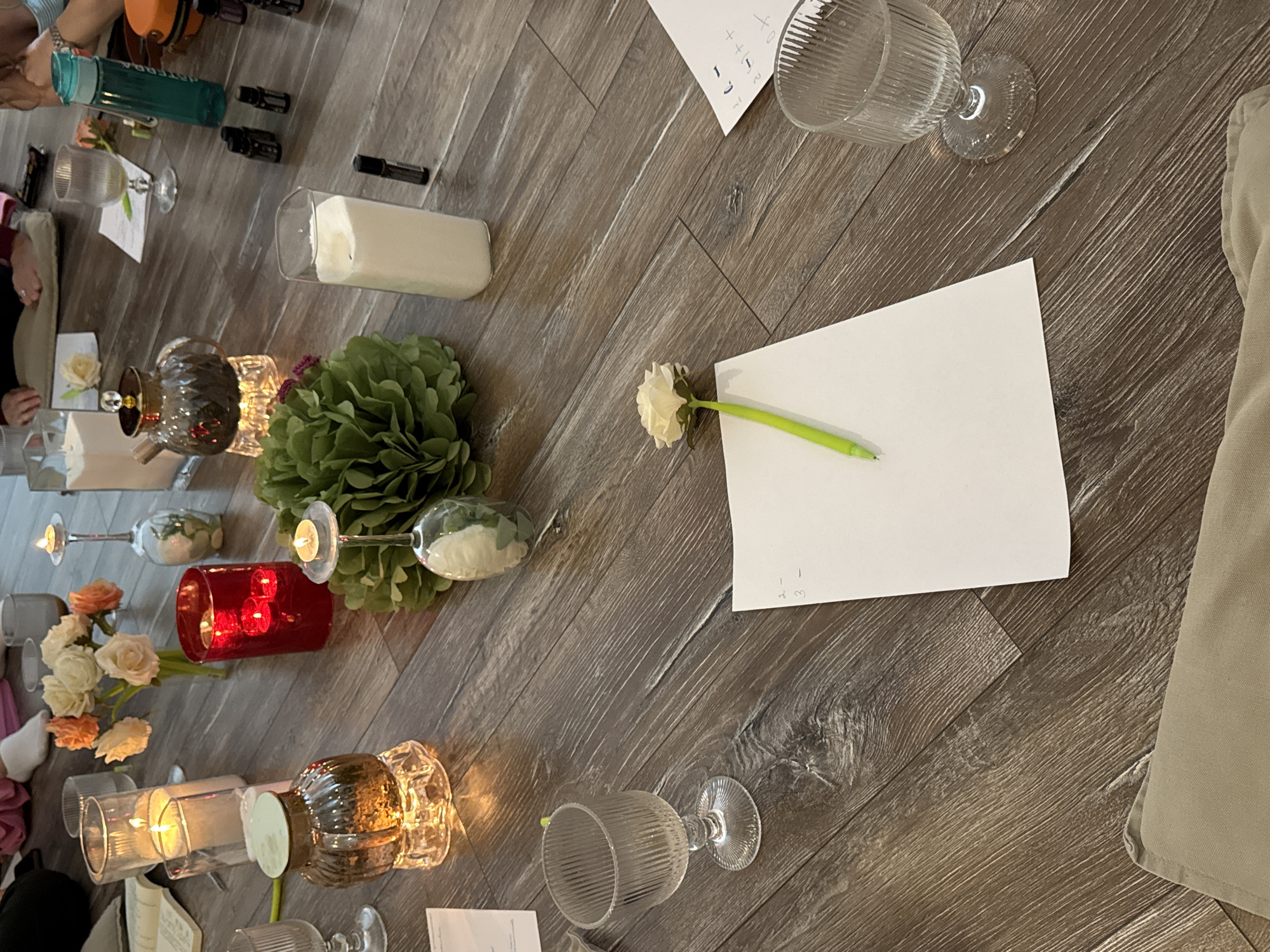 The image shows a collection of items arranged on a dark wood-look floor. The items include candles, flowers, glassware, a green pom-pom decoration, a rectangular white candle, a white sheet of paper, and a green pen. In the upper left of the image, a blue container and several small bottles are visible. A person's hand can also be seen in the upper left, possibly part of an arm extending into the scene. To the right of the flower and pen is a piece of white paper. In the lower right corner, a tan fabric item is situated. The scene is dimly lit by the candles and available lighting in the room. The location is not specified.