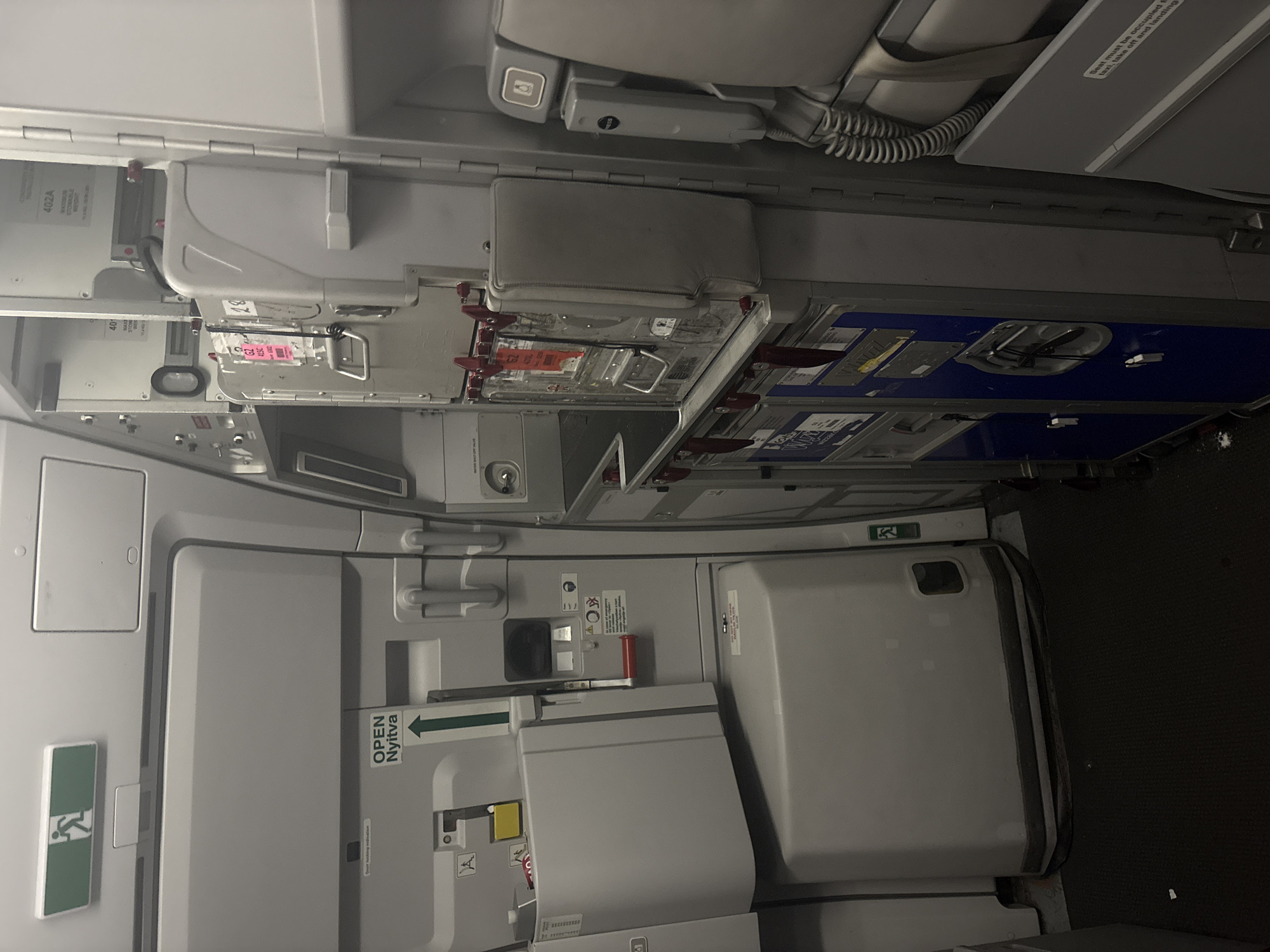 The image depicts the interior of an aircraft, specifically an aisle area with a galley cart. On the left, a large, grey, partially opened door is visible, with an illuminated green exit sign above it. Below the exit sign, there's a sign that reads "OPEN NYITVA" with an upward-pointing arrow. Further down the door, a red handle is present.
To the right of this door, a metal galley cart is positioned, filled with various compartments and latches. Some labels and stickers are affixed to the cart. Above the galley cart, there are panels and storage compartments within the aircraft's structure. On the far right, airplane seats are partially visible, along with a gray telephone handset mounted on the wall. The floor is covered with a dark, textured carpet.
The image does not provide any information about the city of Pitești, Romania, nor does it contain any contextual clues to suggest this location. It is a forensic, matter-of-fact description of the interior of an aircraft.