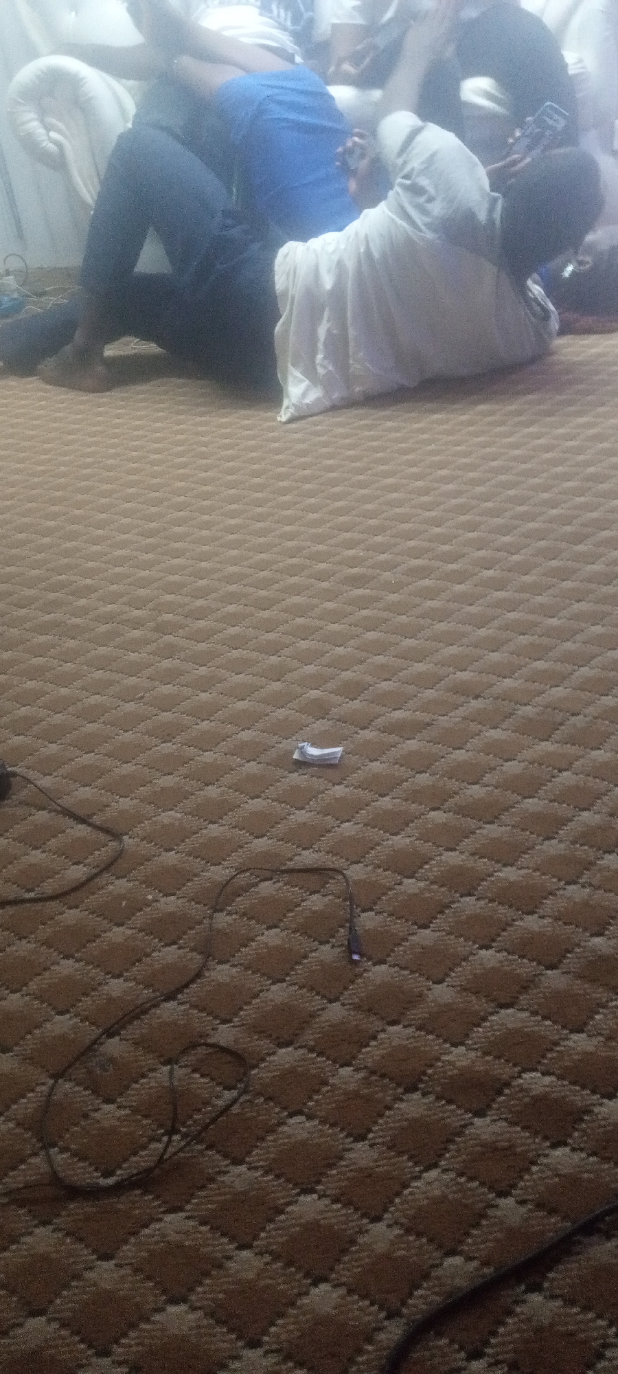 The image shows a carpeted room with several people sitting on a white sofa in the background. One person is wearing a blue shirt and black pants, while another is covered with a white cloth. A black USB cable and a crumpled piece of white paper are on the carpet in the foreground. The location is Unknown, Unknown.