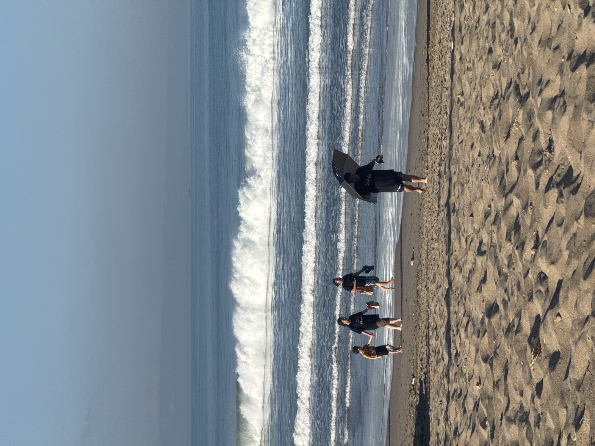 The image depicts a beach scene, possibly in North Kuta, Indonesia. In the background, the ocean rolls with breaking waves. Four people are on the beach. One is standing near the water's edge, holding a boogie board. The other three people are walking near the shoreline. The foreground shows a stretch of sandy beach.