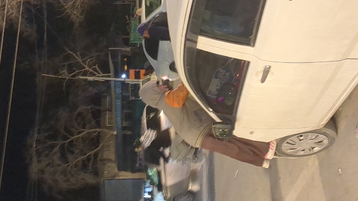 A person wearing a gray hooded sweatshirt and brown pants stands next to a white car on a street at night. The car is parked on the right side of the frame. The person appears to be looking at the car or working on it. Bare trees and streetlights are visible in the background. The image is somewhat dark and grainy, possibly taken in low light conditions.