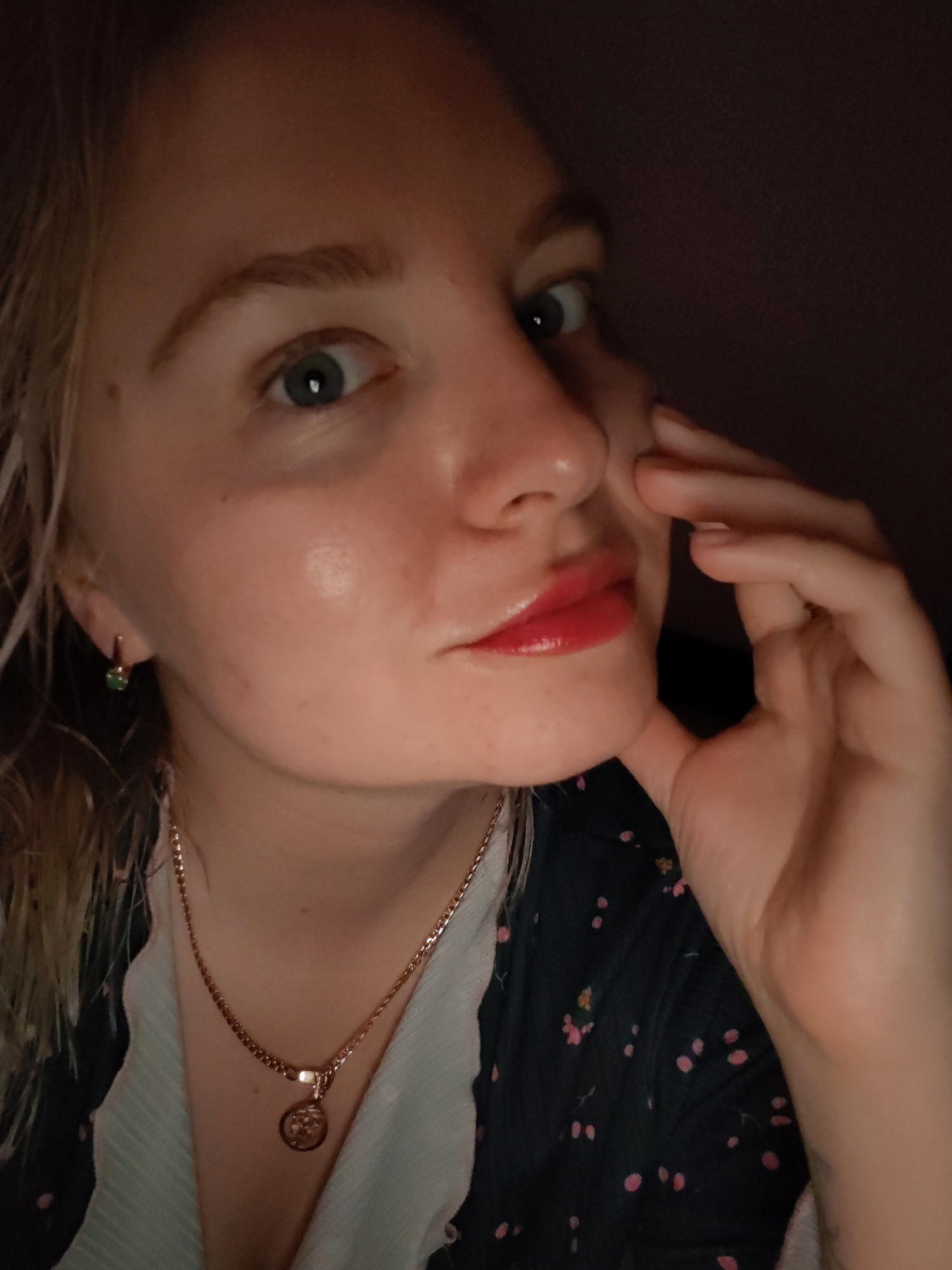 A fair-skinned woman with blonde hair is posing indoors. She wears red lipstick, a gold chain necklace with a pendant, and earrings with green stones. She has on a dark-colored shirt with a small pink floral pattern. Her right hand is touching her face. The background is dark. There is no information to suggest that the image was taken in Izhevsk, Russia.