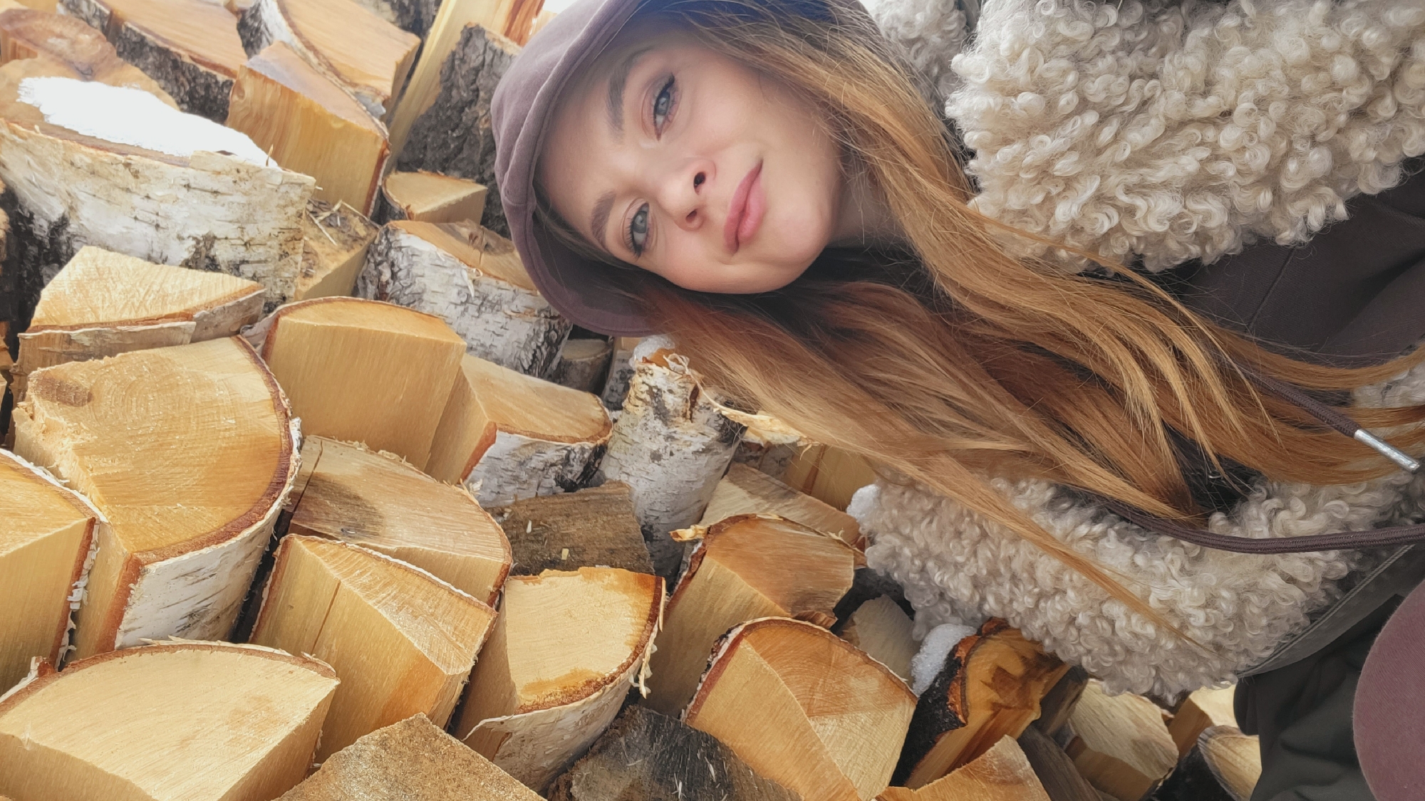 A woman wearing a brown hat and fur-lined coat reclines among a pile of firewood in Зипуново, Russia. The firewood is comprised of birch logs. The woman has long, reddish-brown hair and is smiling.