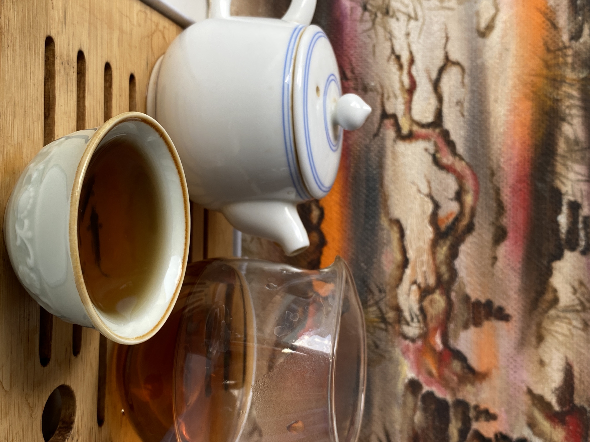 The image shows a teacup filled with tea, a white teapot with blue lines, and a glass filled with tea, all resting on a wooden surface with vertical slots. Behind these items is a colorful abstract painting or textile. The location is not discernible from the image.
