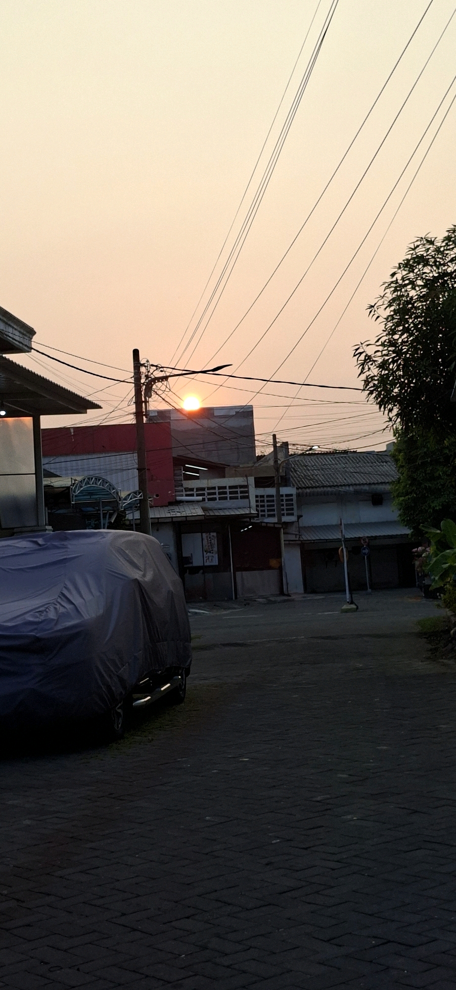 The image captures a sunset over a residential street in Surabaya, Indonesia. The sky is a soft gradient of orange and yellow, with the sun peeking over the rooftops of buildings in the background. Numerous power lines crisscross the sky, creating a layered effect. In the foreground, a dark-colored car is covered with a tarp. The street itself is paved with interlocking bricks. Buildings line the street, some with visible windows and awnings, suggesting a commercial or mixed-use area. A tree with dense foliage is visible on the right side of the frame. The overall impression is a quiet, everyday scene at dusk in an Indonesian city.