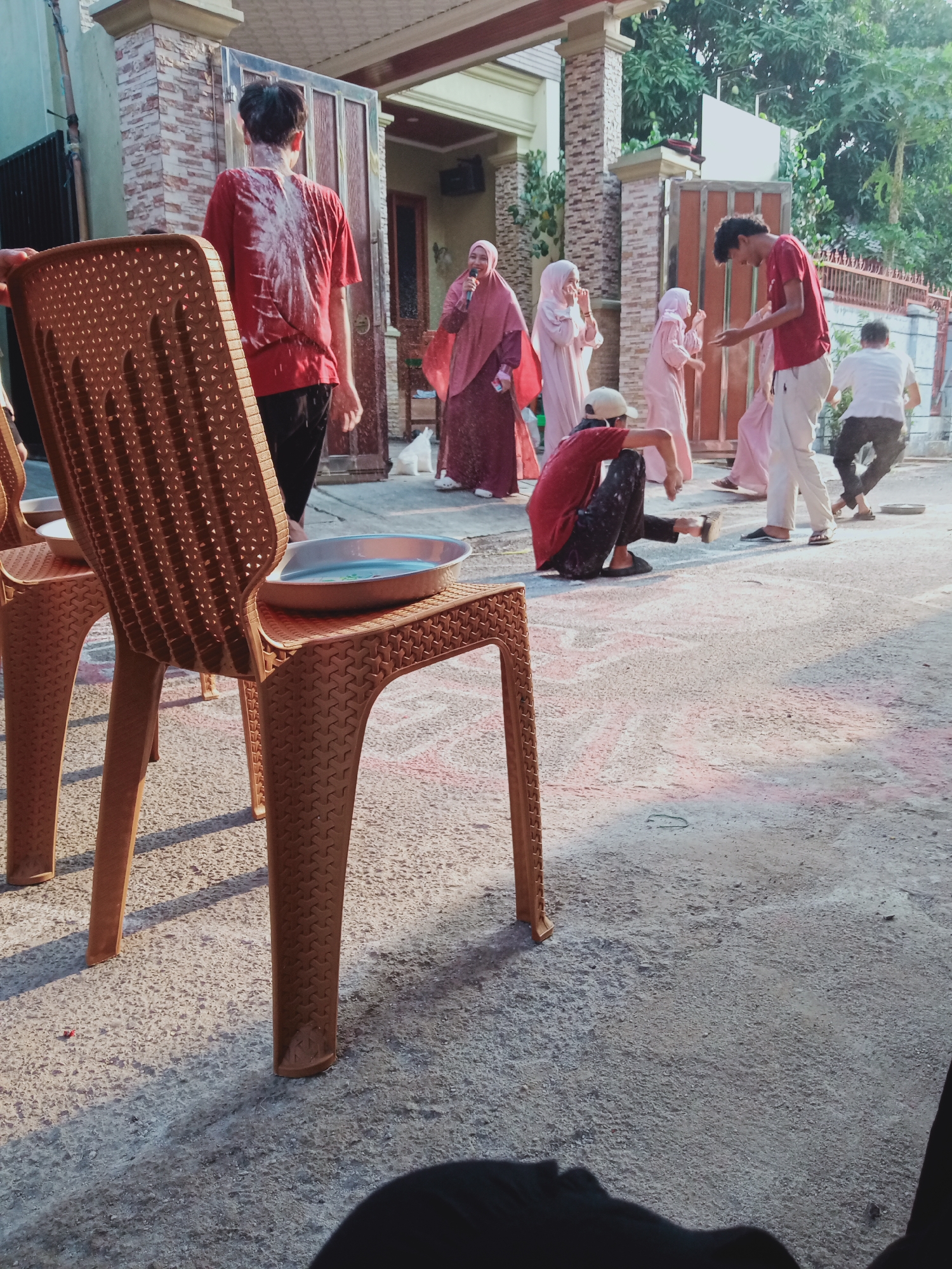 In East Jakarta, Indonesia, an outdoor scene depicts a group of young people engaged in a festive activity, likely a water-based game given the presence of a basin of water on a chair. The setting appears to be a residential area, with a house featuring a textured brick facade and a large metal gate in the background. Several individuals are captured in motion, suggesting a playful and energetic atmosphere.
In the foreground, a brown plastic chair occupies the left side of the frame, with a metal basin filled with water placed on its seat. Behind the chair, a young person in a red t-shirt and black pants is the central focus, with water or some white liquid splashed on their back. To their right, several other young people are visible. One individual, also in a red shirt, is crouched on the ground, facing away from the camera, while others in pink hijabs and dresses are standing, looking towards the person being splashed. Another young person in a red t-shirt and white pants is standing and gesturing, possibly participating in or observing the activity. Further in the background, a person in a white t-shirt and black pants is running, and another figure in a white t-shirt is further to the right. The ground is a paved surface, possibly a driveway or courtyard, with what appears to be colored chalk markings scattered across it. The overall lighting suggests it is daytime, with sunlight casting shadows.