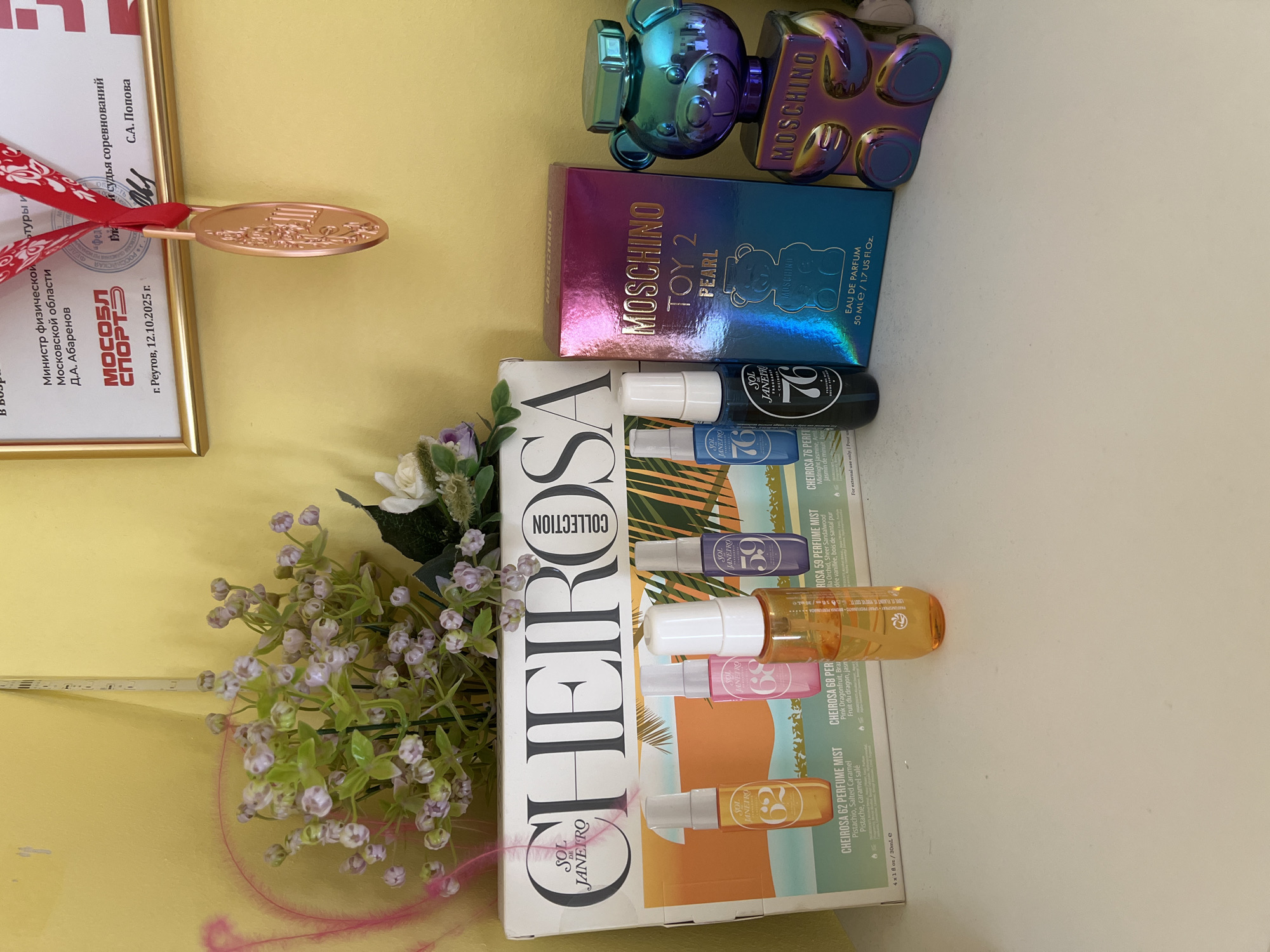 A medium shot captures a collection of perfumes and decorative items displayed on a white surface against a yellow wall. On the left, a box for "Sol de Janeiro Cheirosa Collection" holds four mini perfume mist bottles labeled "62", "68", "59", and "76". Three of these bottles are visible: "62" (yellow), "68" (pink), and "59" (blue), with the "76" bottle also visible on the far right of the box. In the center, a box for "Moschino Toy 2 Pearl" eau de parfum sits next to a unique, iridescent bear-shaped perfume bottle also by Moschino. To the left of the Sol de Janeiro box, a bouquet of artificial light purple flowers with green leaves is arranged, with a pink decorative feather to its left. Above the perfumes, a framed certificate and a gold medal hang on the yellow wall. The certificate, with red and white lettering, mentions "Министр физической культуры Московской области" and "Реутов, 12.10.2025 г.". The medal is gold and oval-shaped, suspended by a red ribbon. The scene is set indoors.