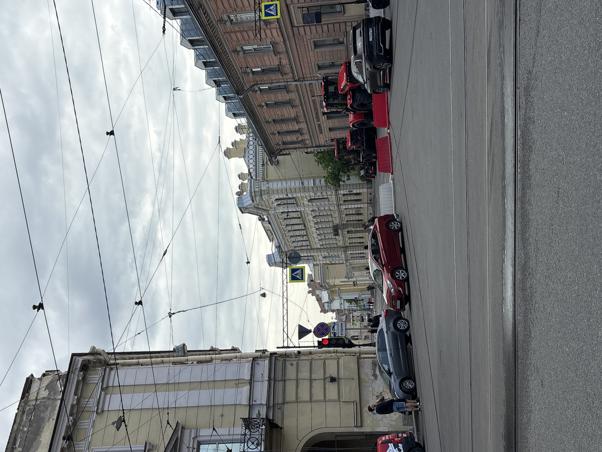 The image depicts a street scene in Saint Petersburg, Russia. The view is taken from a low angle, looking down a city street. The sky is overcast. The street is lined with buildings on both sides, and above, there are a network of electrical or tram lines.
On the right side of the street, several vehicles are parked in a row including a black SUV, several red tractors with red and white barricades, and a red sedan.
On the left side of the street, there is a gray car and two people are standing on the sidewalk. There are also visible traffic signs and a traffic light.