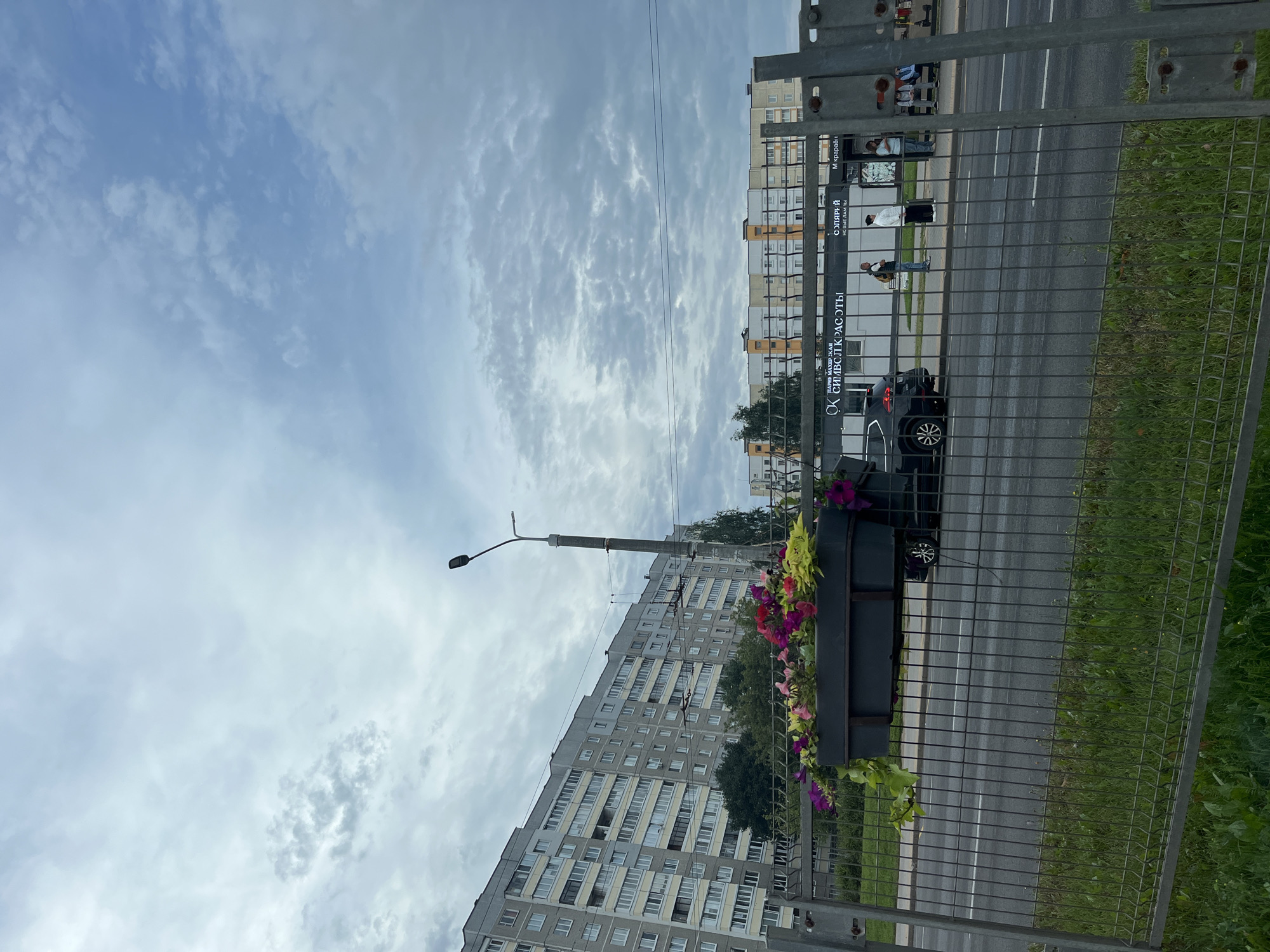 The image depicts a street scene in Minsk, Belarus, with a grey sky filled with scattered clouds. In the foreground, a metal fence separates the viewer from the street. Beyond the fence, a black car is parked next to a planter box overflowing with colorful flowers, including pink, purple, and yellow blossoms, and trailing green foliage. The car is positioned on the side of a road.
Further down the street, a light-colored apartment building with numerous windows stretches across the left side of the frame, dominating the background. To the right of the apartment building, a row of businesses with awnings and signage is visible. One sign reads "ОК СИМВОЛ КРАСОТЫ" (OK Symbol of Beauty) and another "ОЛЯЙЯЙ" (Olyayyay). Several people are standing on a sidewalk in front of these establishments. One woman in a white top and dark skirt is holding a black bag. Another person wearing a light blue shirt and dark pants stands near a bus stop shelter. Two people, one in a light blue shirt and the other in a white shirt, are seated on a bench within the shelter. A street lamp stands tall near the apartment building. The overall environment appears to be an urban residential and commercial area.