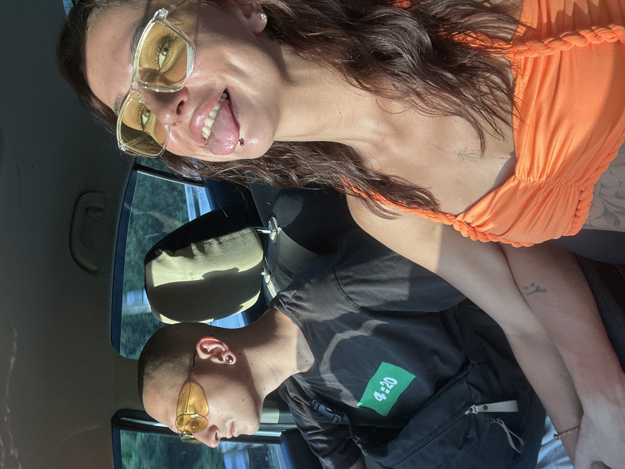 The image shows two individuals in a car on a sunny day, possibly on Koh Samui, Thailand, given the lush greenery visible outside. The person in the foreground, on the right, is a woman with dark hair, wearing an orange top and yellow-tinted glasses. She is sticking her tongue out and has a lip piercing. Her right arm is visible, showing a tattoo on her forearm. The person in the background, on the left, is a man wearing a black t-shirt with a green "4:20" logo and yellow-tinted glasses. He is looking out the window. Both individuals appear to be enjoying a car ride, with the sunlight casting shadows within the vehicle. The car's interior, including the seats and headrests, is visible, indicating a typical passenger vehicle setting. The overall atmosphere is casual and relaxed.